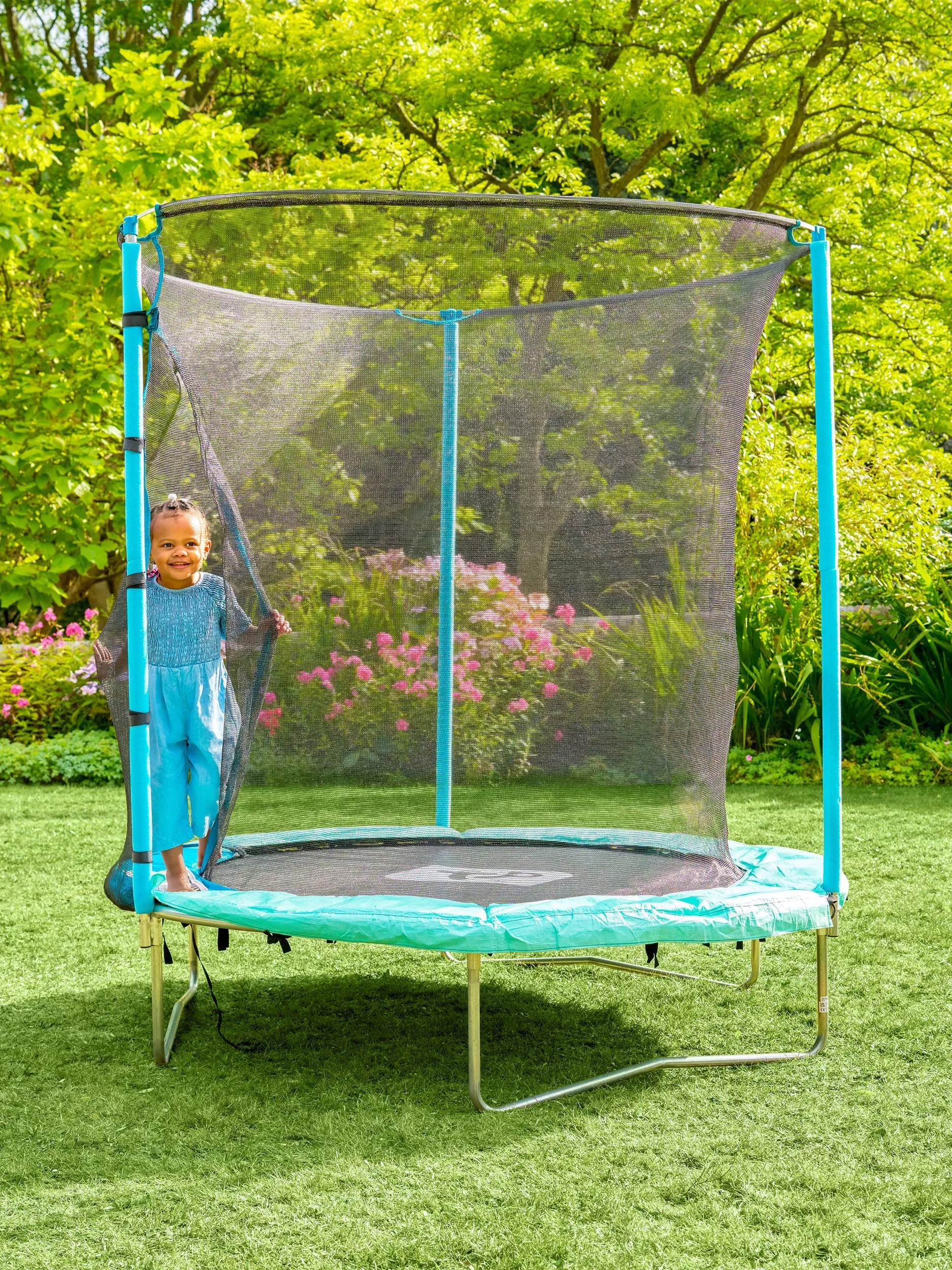 A young child stands on a trampoline with a safety net in a garden, surrounded by lush greenery and flowers.