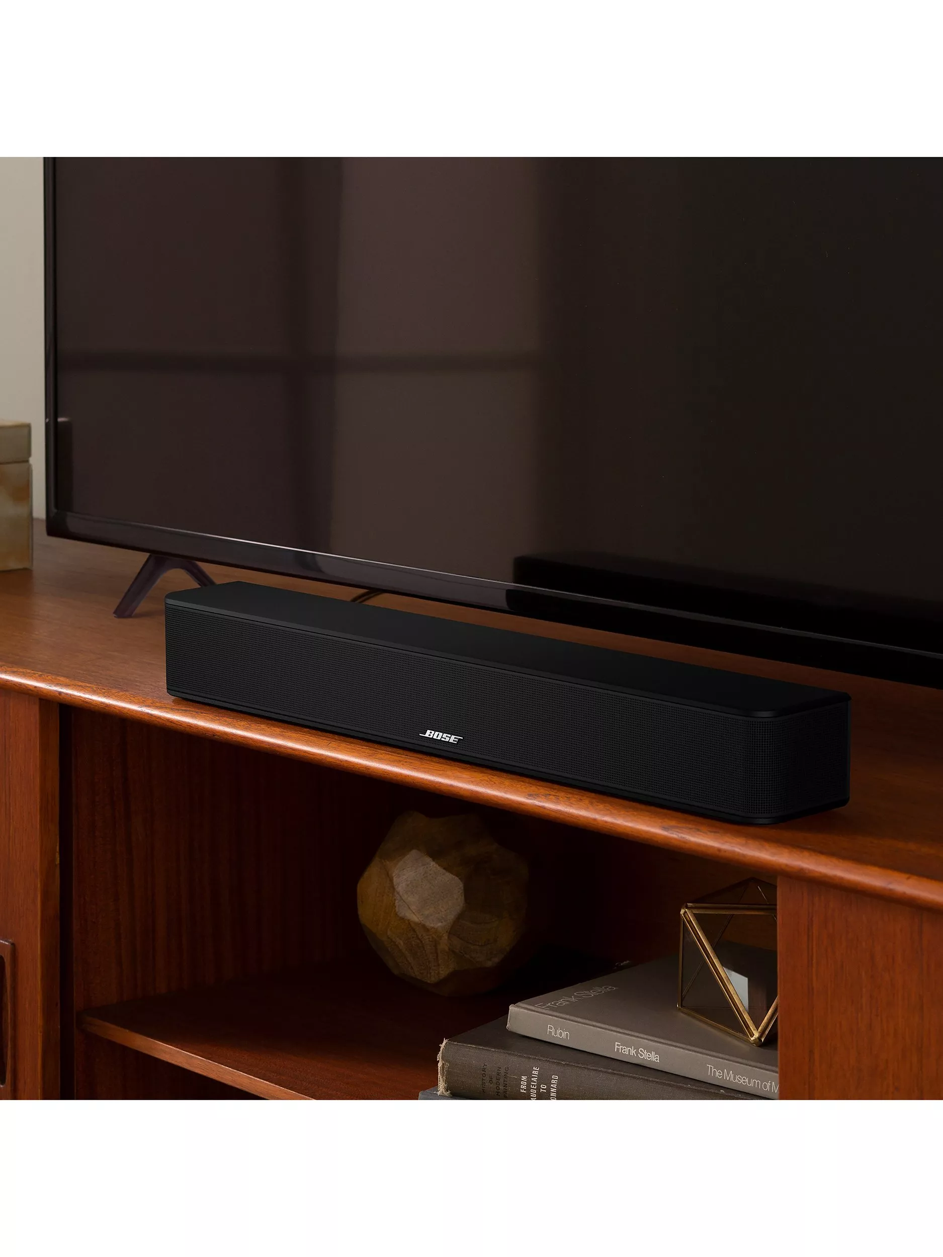 A sleek black soundbar sits on a wooden shelf beneath a large TV, with books and a decorative item nearby.