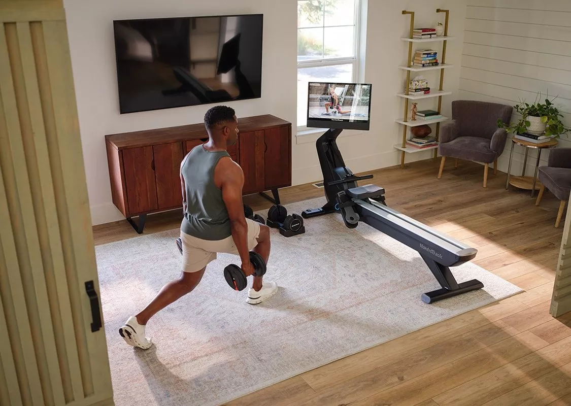 Man doing dumbbell lunges in a home gym living room with rowing machine and wall-mounted TV for workouts