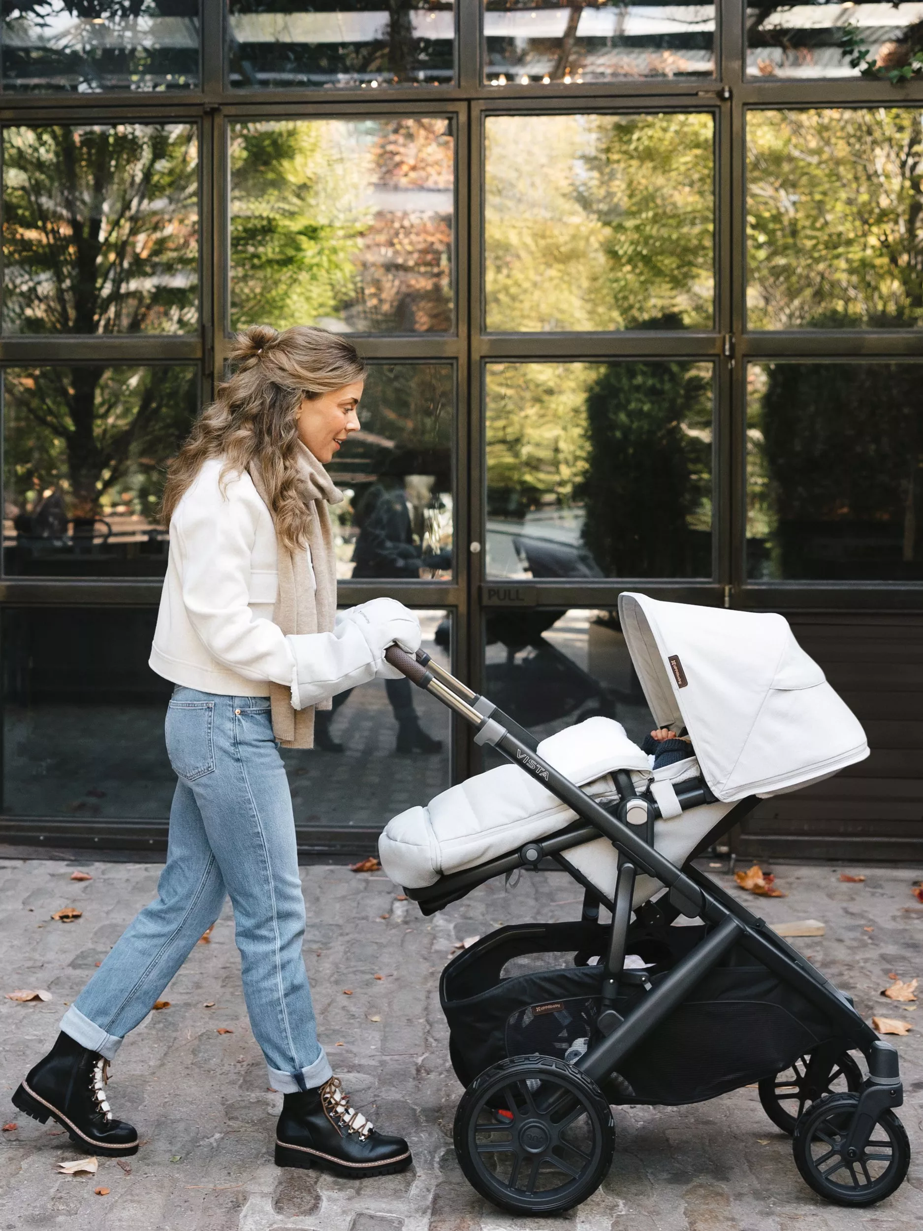 Woman pushing a modern baby pram on a city pavement, autumn leaves and glass-fronted building behind.