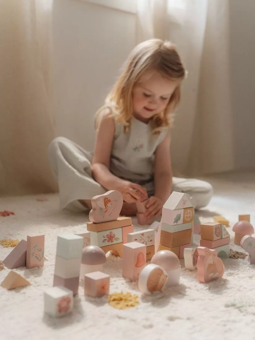 Toddler girl playing with pastel wooden blocks on a soft rug in a sunlit nursery, building a toy house.