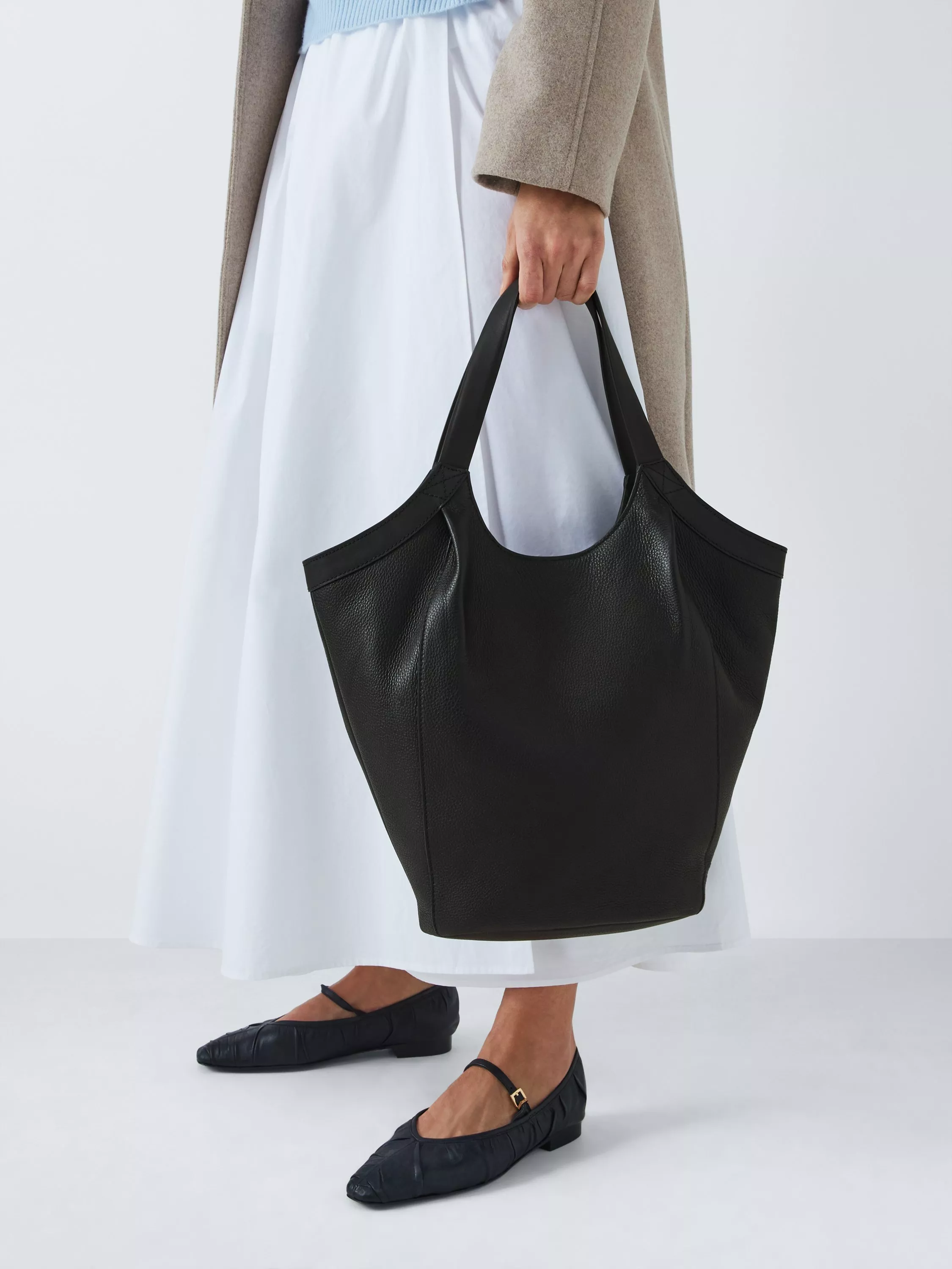 A person holds a large black leather tote bag. They're wearing a white skirt, beige coat, and black flats.