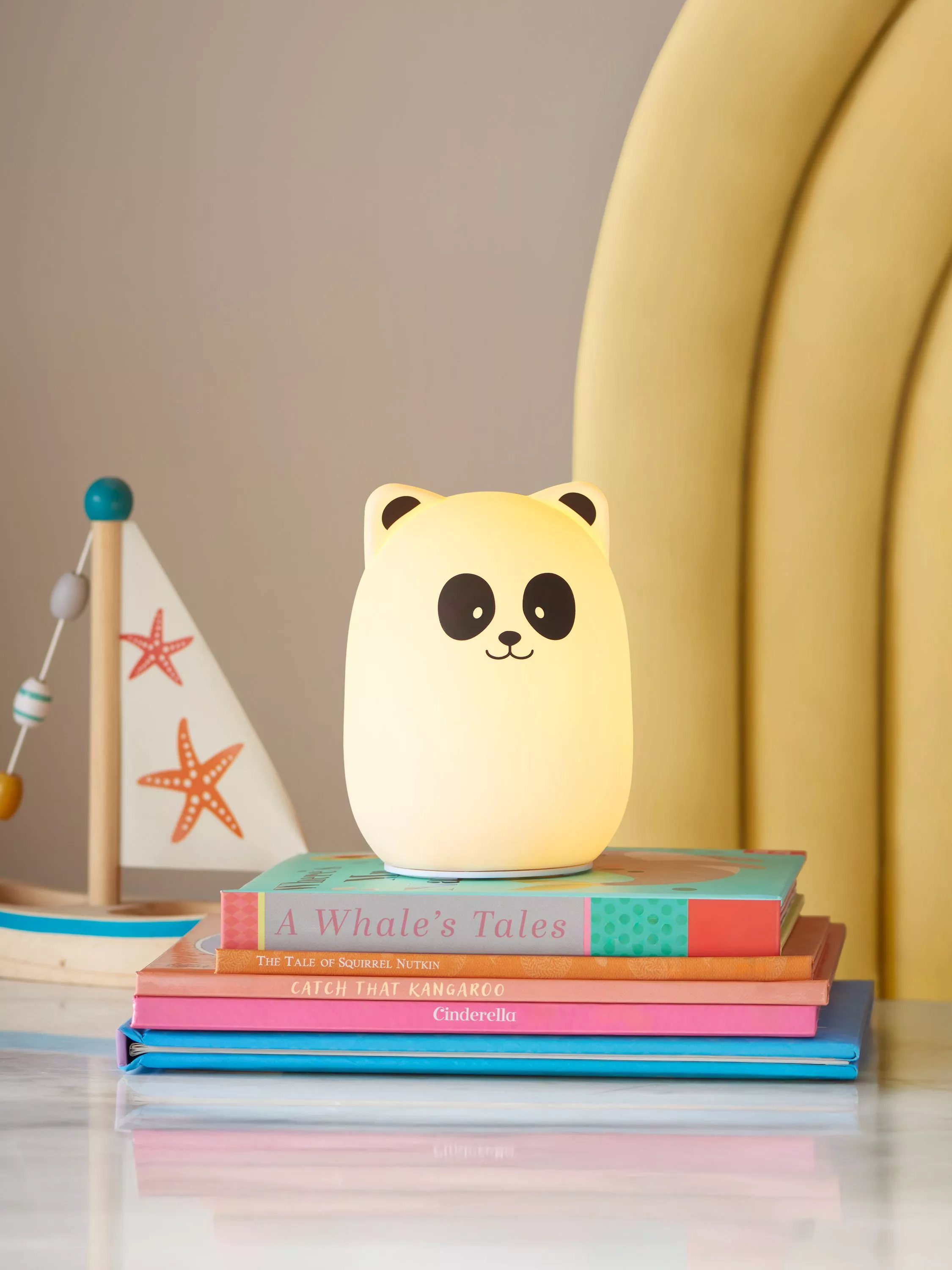 Cute panda night light on stacked children’s books beside toy sailboat, cosy nursery bedside decor and lighting