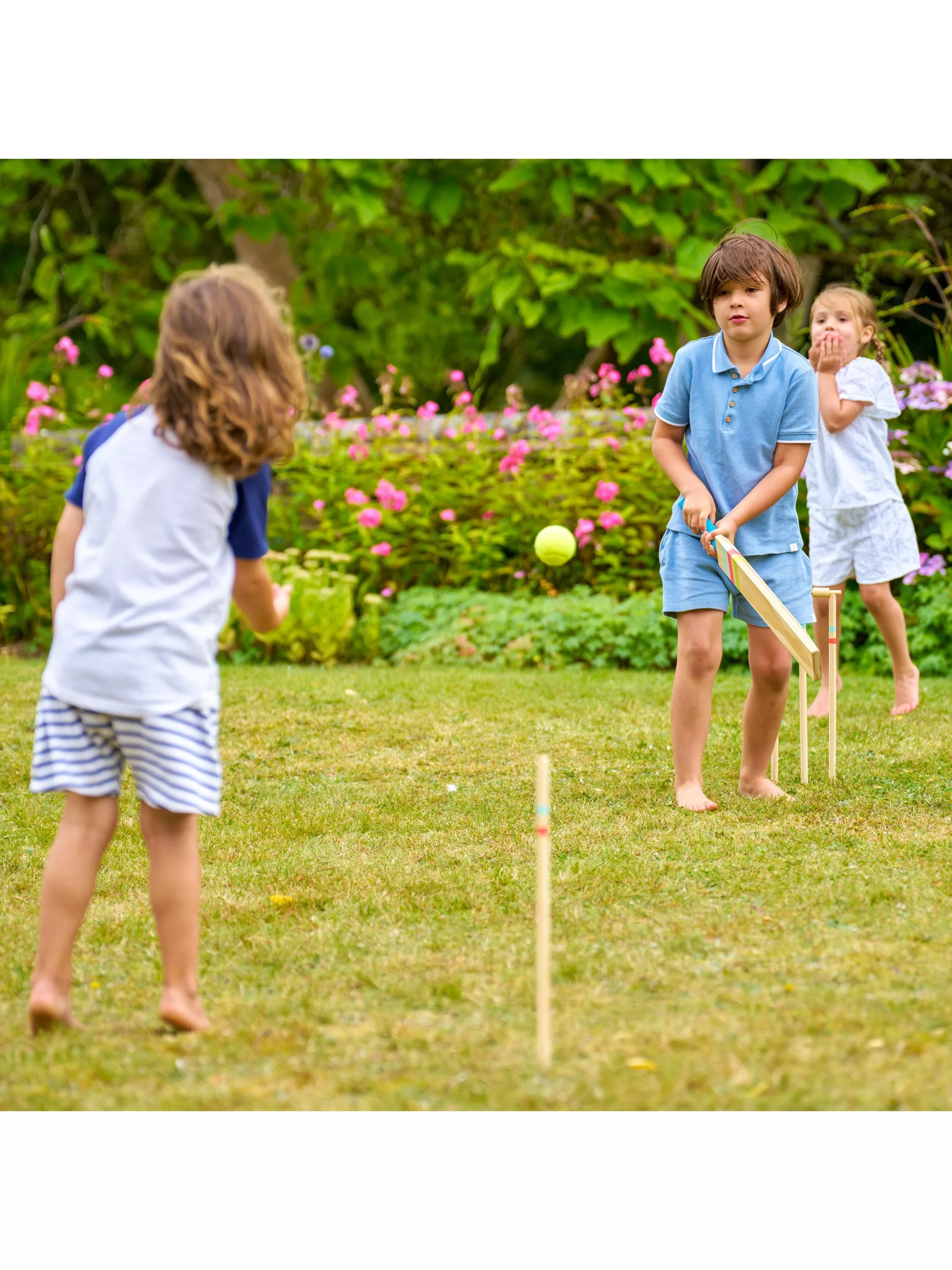 Children playing garden cricket on a summer lawn, one batting as the ball is bowled, flowers behind
