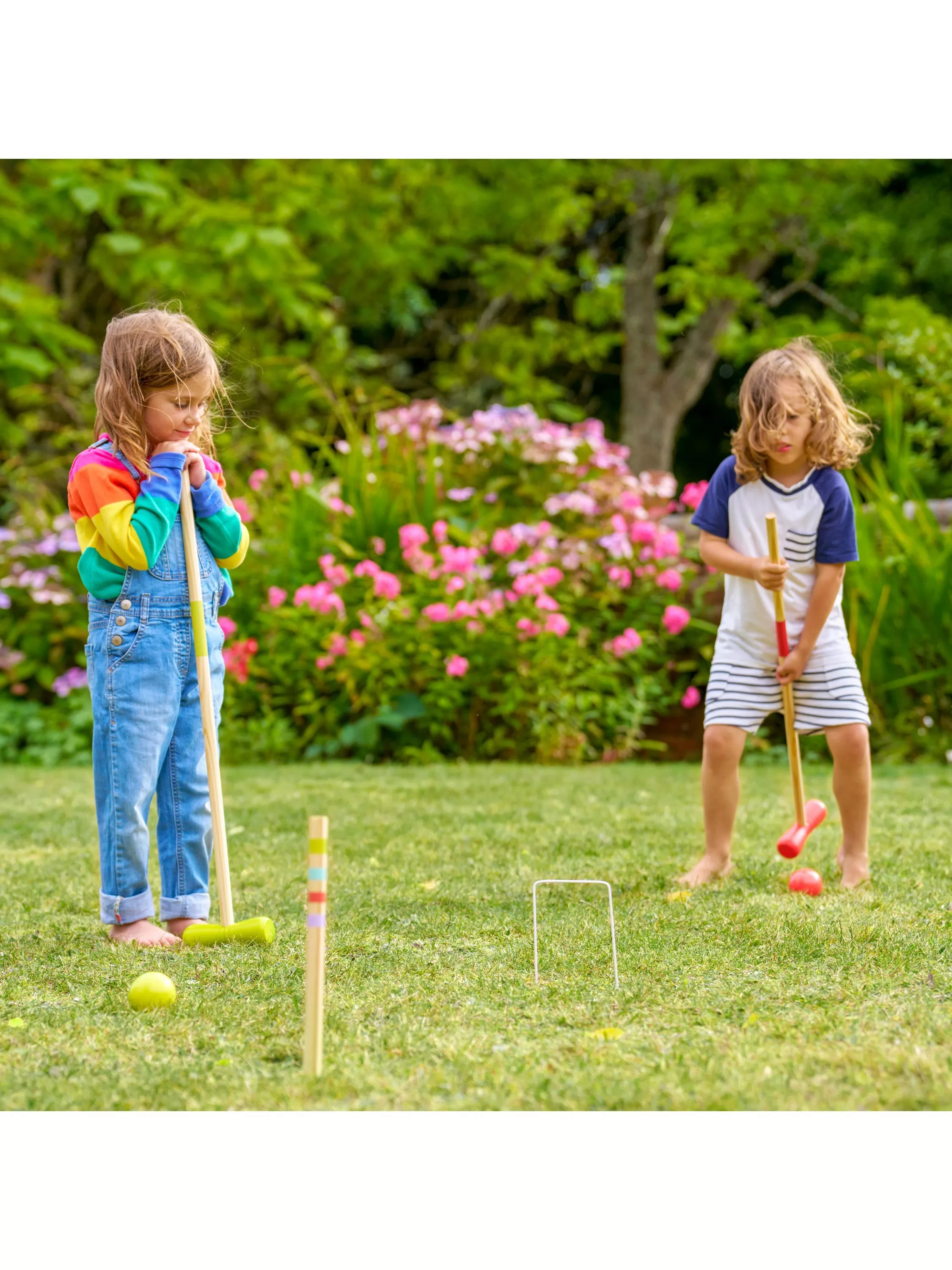 Two children play outdoor croquet on a grassy lawn surrounded by blooming flowers and lush greenery.