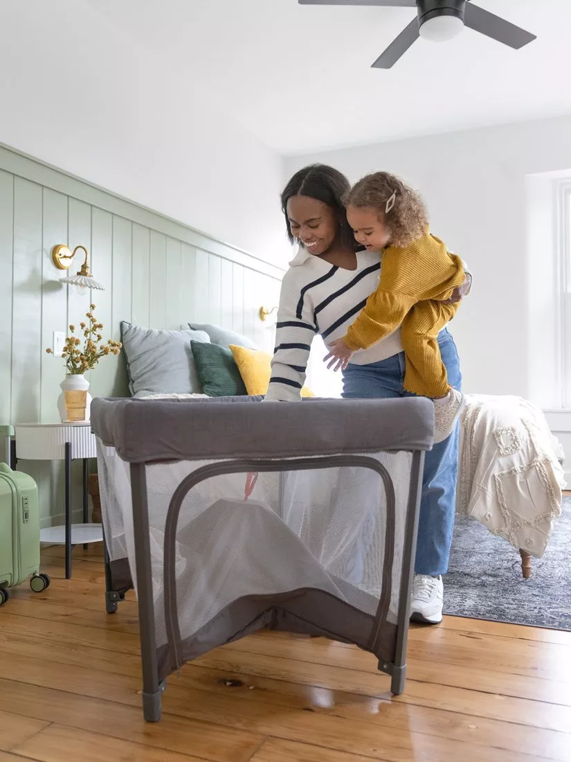 Mother holding toddler beside a portable travel cot in a bright bedroom, setting up baby sleep space at home