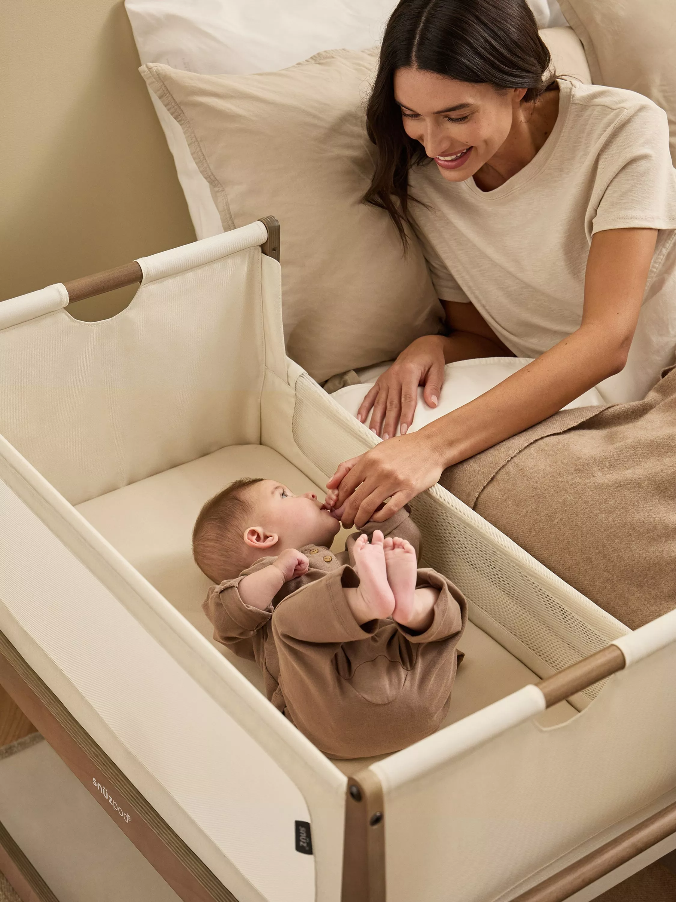 Mother soothing baby in bedside bassinet next to bed, cosy nursery sleep setup with neutral bedding and wood frame