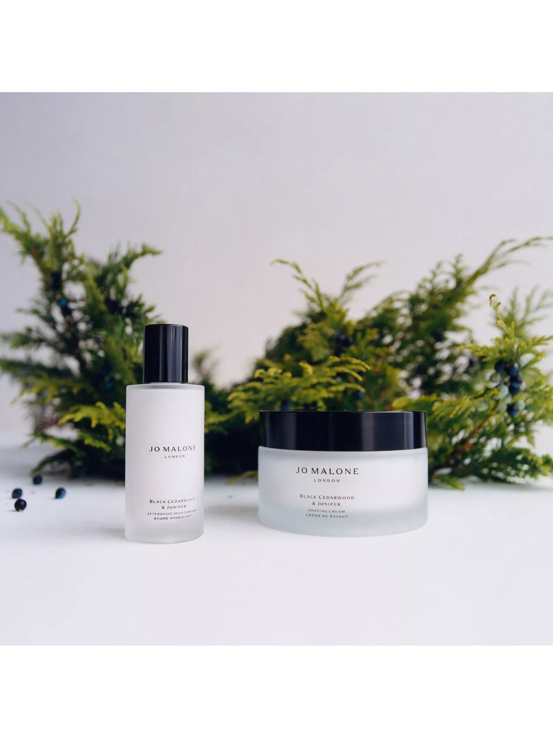 Jo Malone London Black Cedarwood & Juniper aftershave moisturiser and shaving cream, with greenery backdrop.