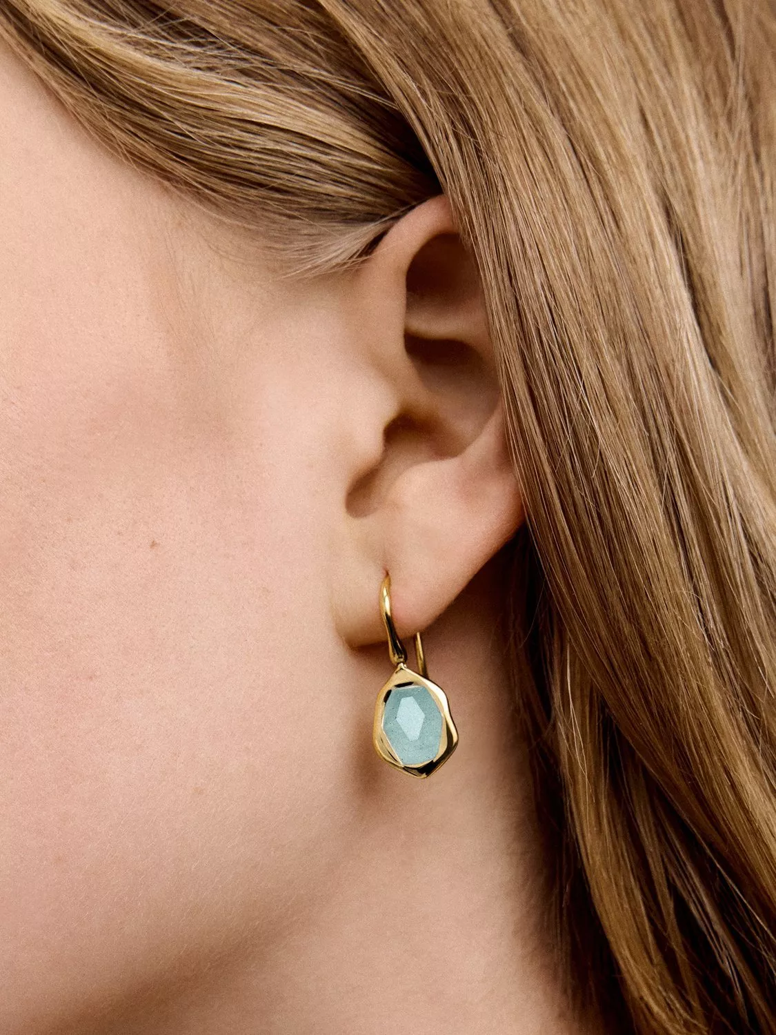 Close-up of an ear wearing a gold earring with a faceted blue gemstone, framed by light brown hair.