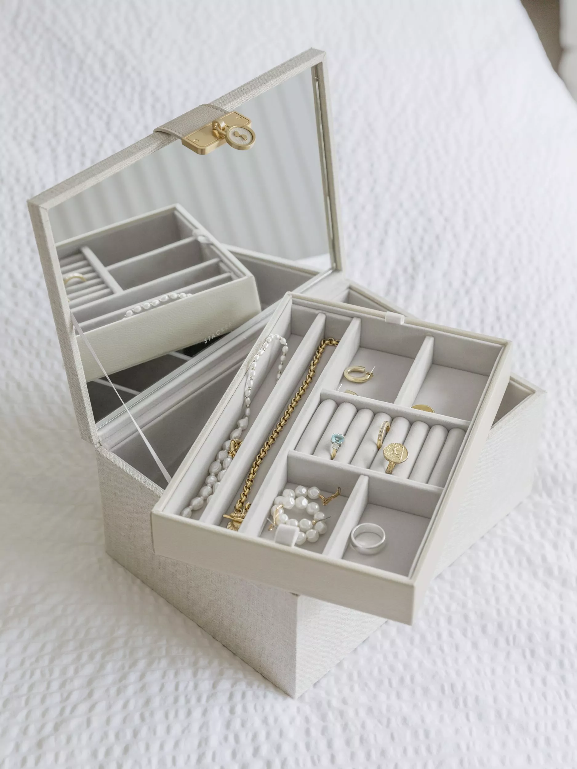 Open mirrored jewellery box with organised trays holding rings, earrings, pearls and gold chain on white bedding