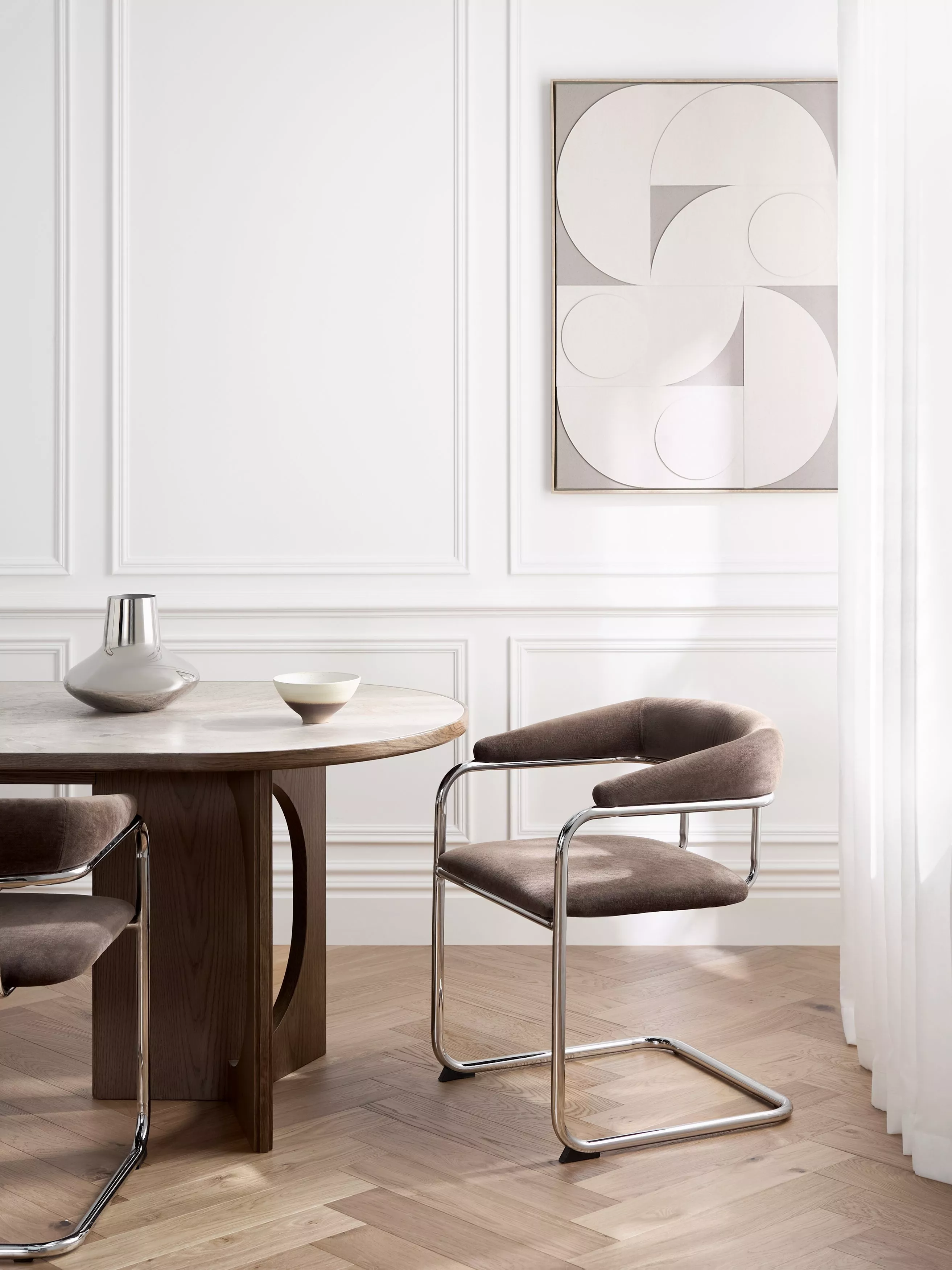 Modern dining room with round wooden table, chrome cantilever chairs and neutral abstract wall art in bright light