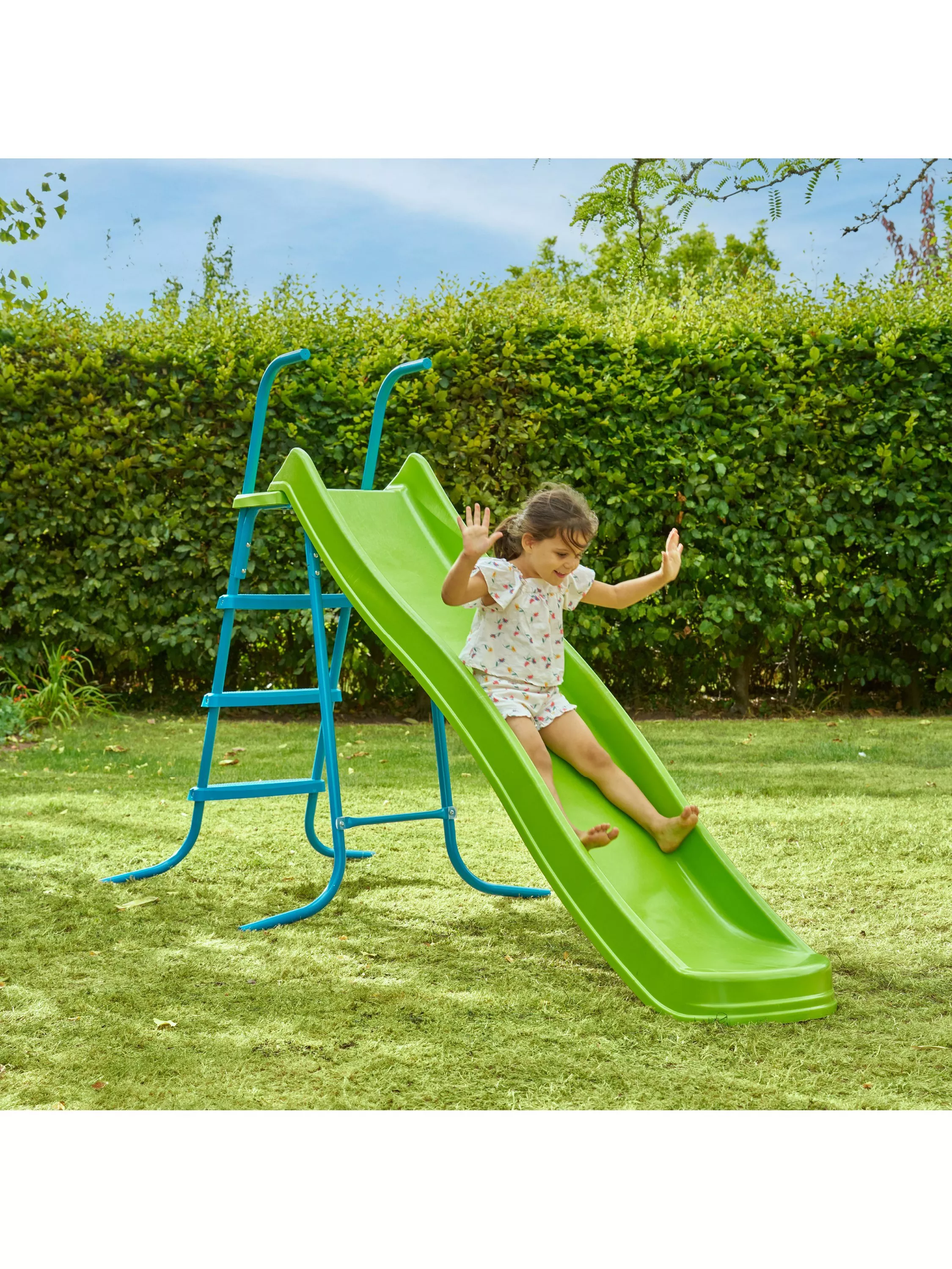 Child joyfully sliding down a bright green slide in a sunny garden, surrounded by lush greenery.