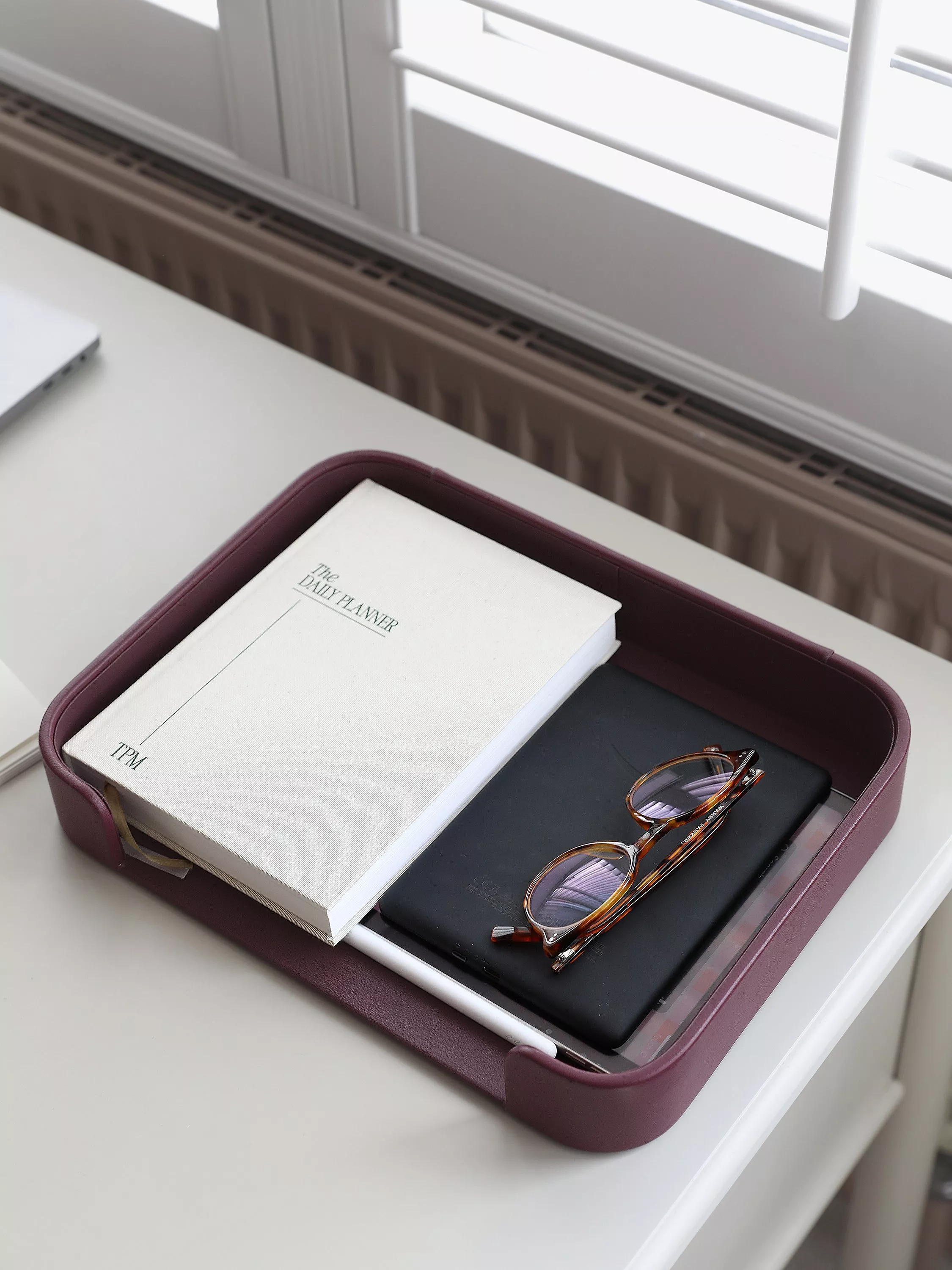 Leather desk organiser tray with daily planner, tablet and tortoiseshell glasses by a window in home office.