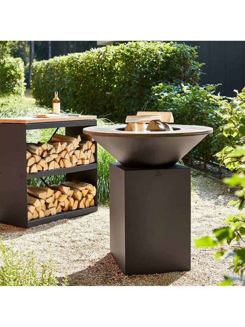 Modern outdoor fire bowl on stand beside wood storage table, garden patio setting with stacked logs and bottle