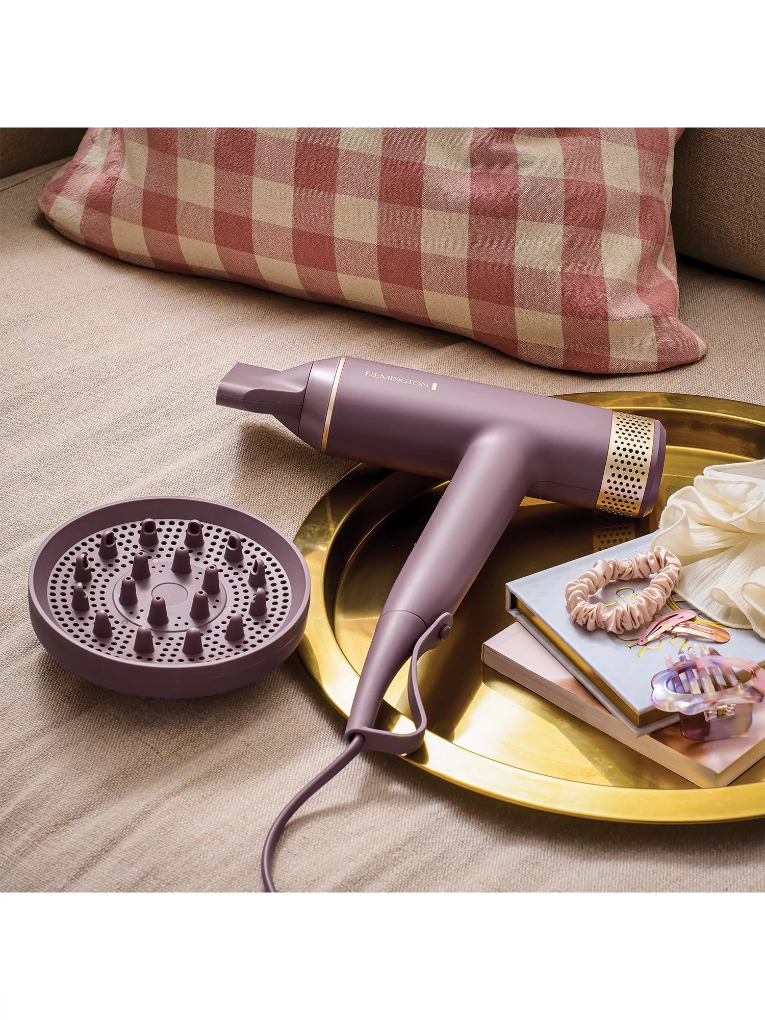 Remington purple hair dryer with diffuser attachment on gold tray beside hair clips and books on sofa