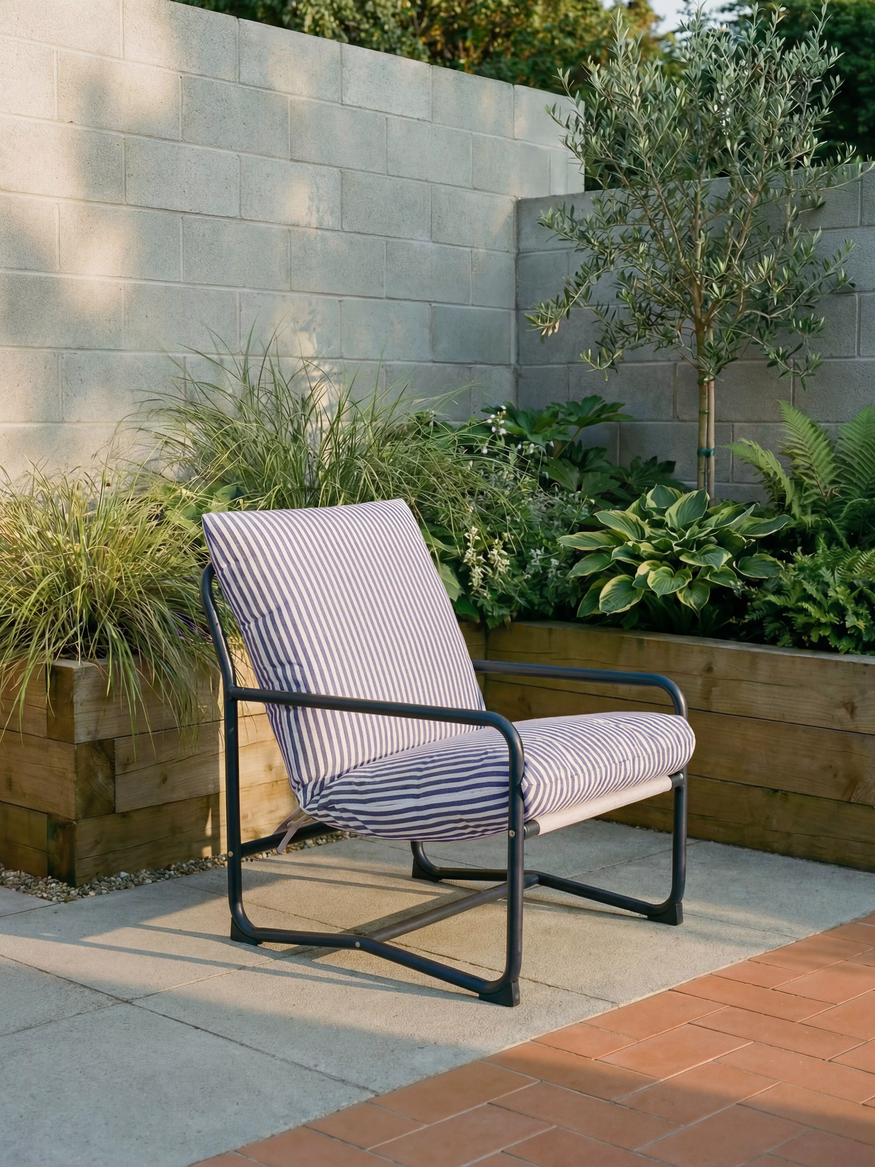 Striped outdoor lounge chair on modern patio, beside raised planter with olive tree and lush garden plants