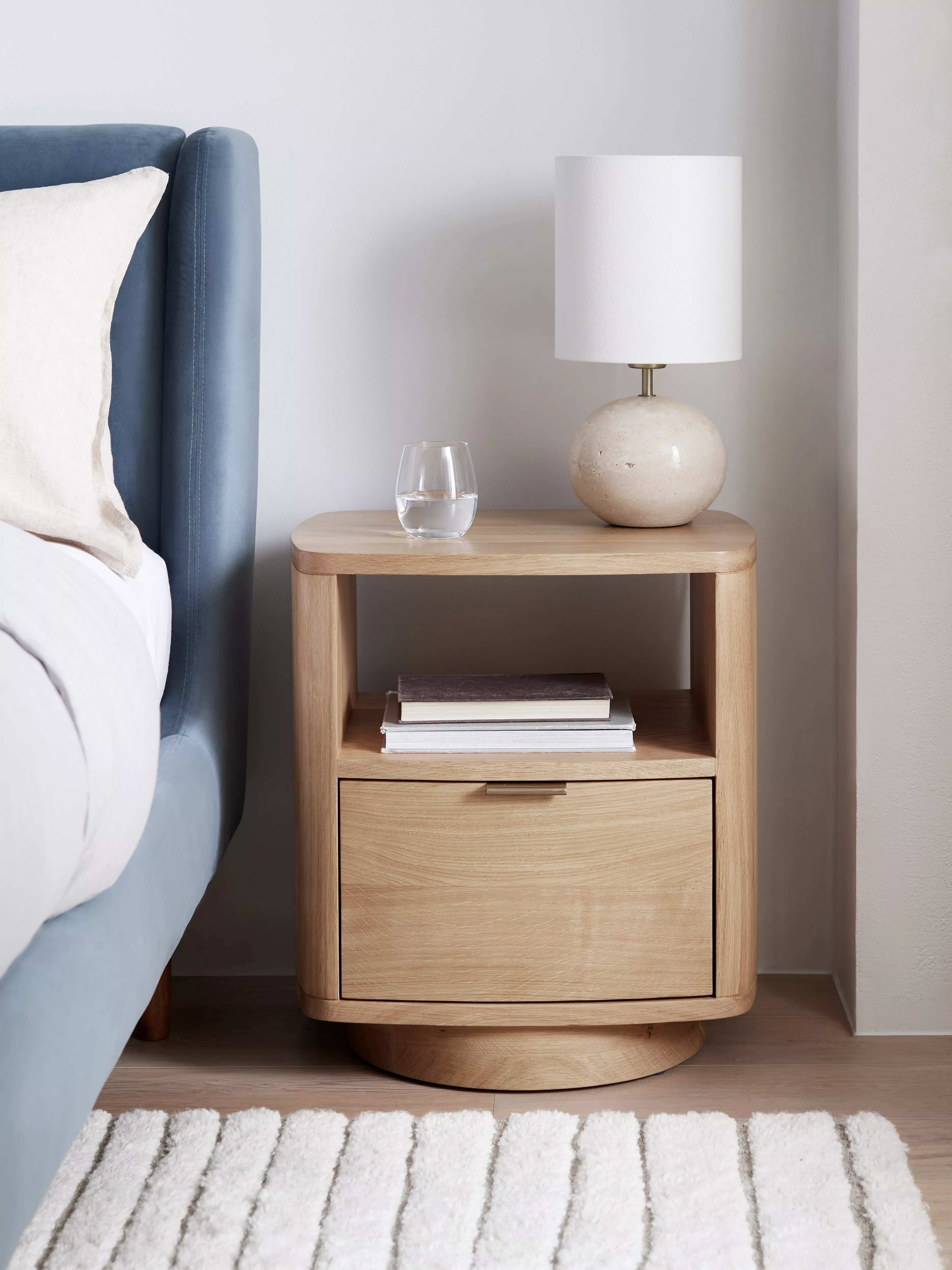 Mid-century wooden bedside table beside upholstered bed, with stacked books and soft morning light on tiled floor