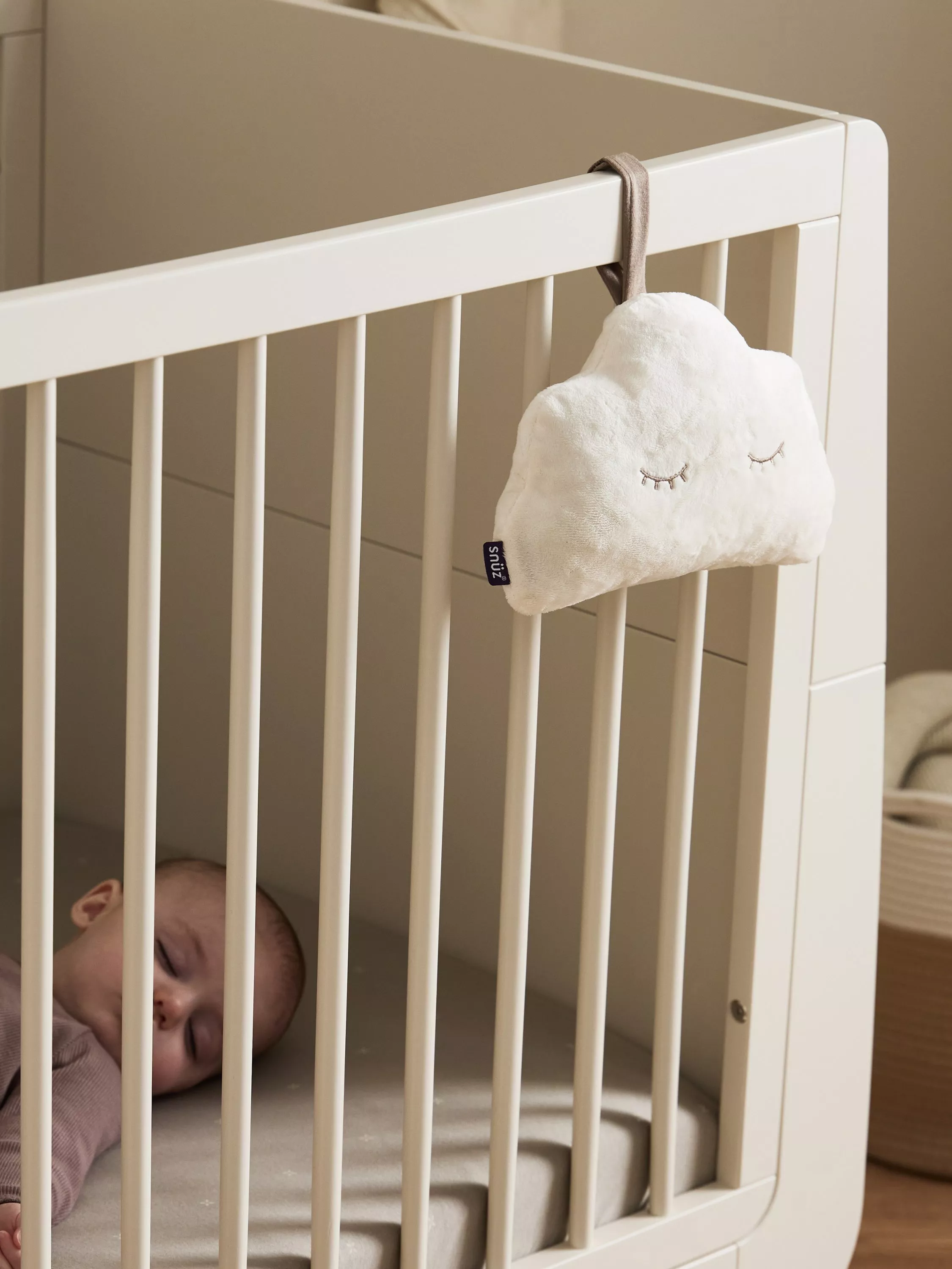 White baby cot with cloud-shaped plush toy hanging on the rail in a calm beige nursery setting