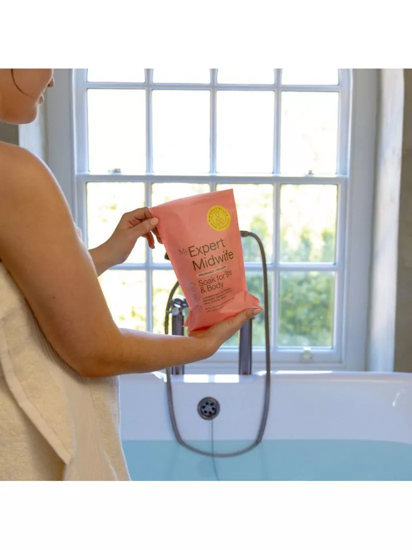 Person holding My Expert Midwife bath salts beside a filled bathtub in a bright bathroom with window backdrop