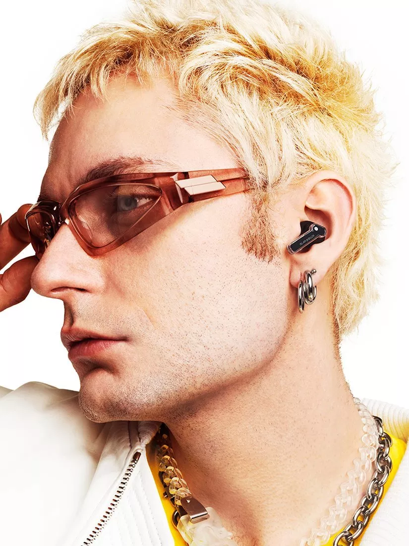 Close-up profile of man with bleached hair, tinted sunglasses, wireless earbud and hoop earrings on white background