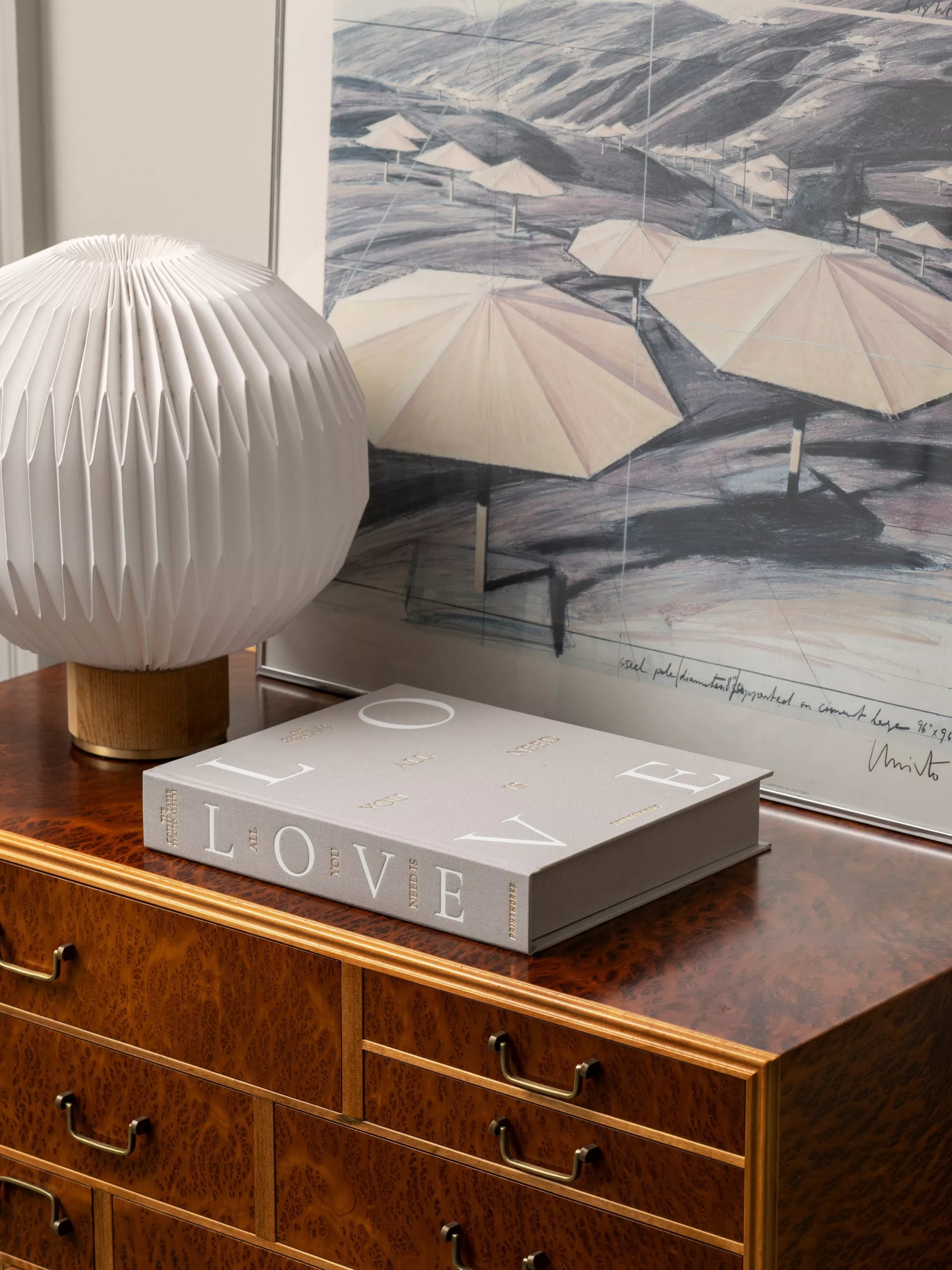 White pleated table lamp beside LOVE coffee table book on wooden sideboard with framed beach umbrella art print.