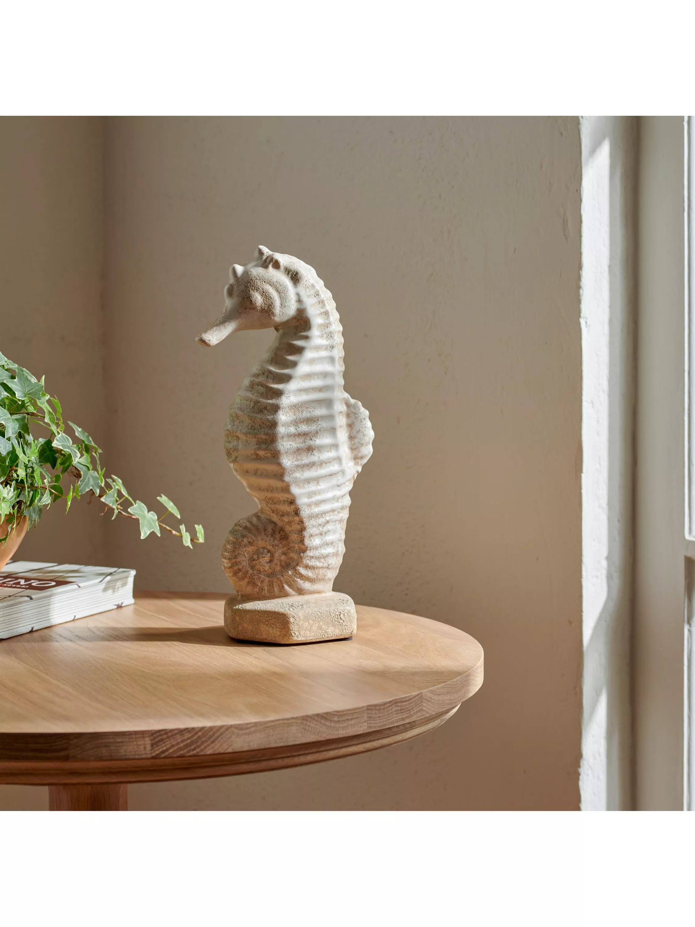 Stone-effect seahorse ornament on a wooden side table by a window, coastal home décor with plant and books