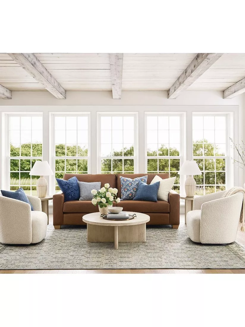 Bright modern living room with brown sofa, boucle armchairs, round coffee table and large windows, neutral decor