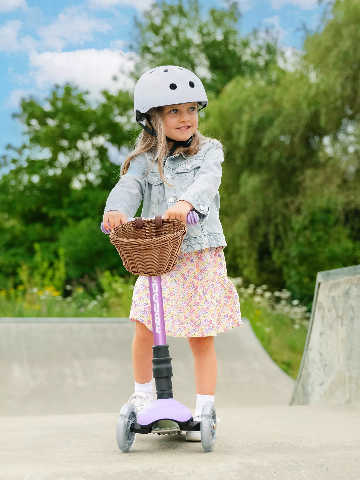 A child wearing a helmet rides a purple scooter with a wicker basket at a skate park, surrounded by greenery.