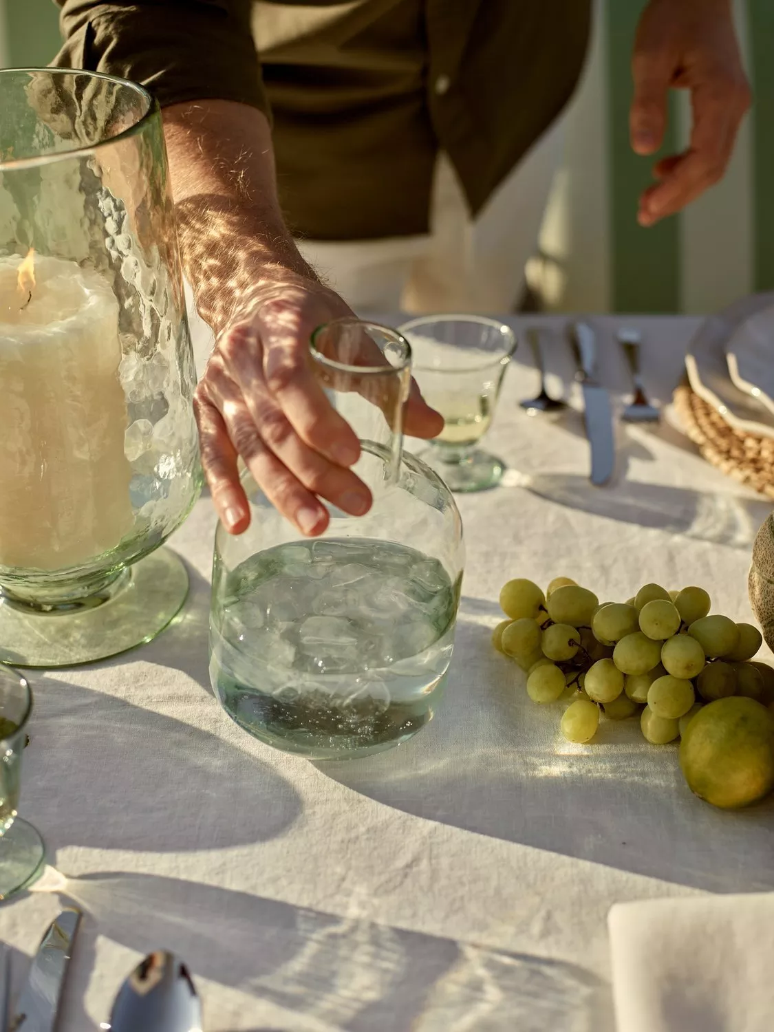 Hand reaching for glass water carafe on sunlit outdoor dining table with grapes, lemon, cutlery and candle jar