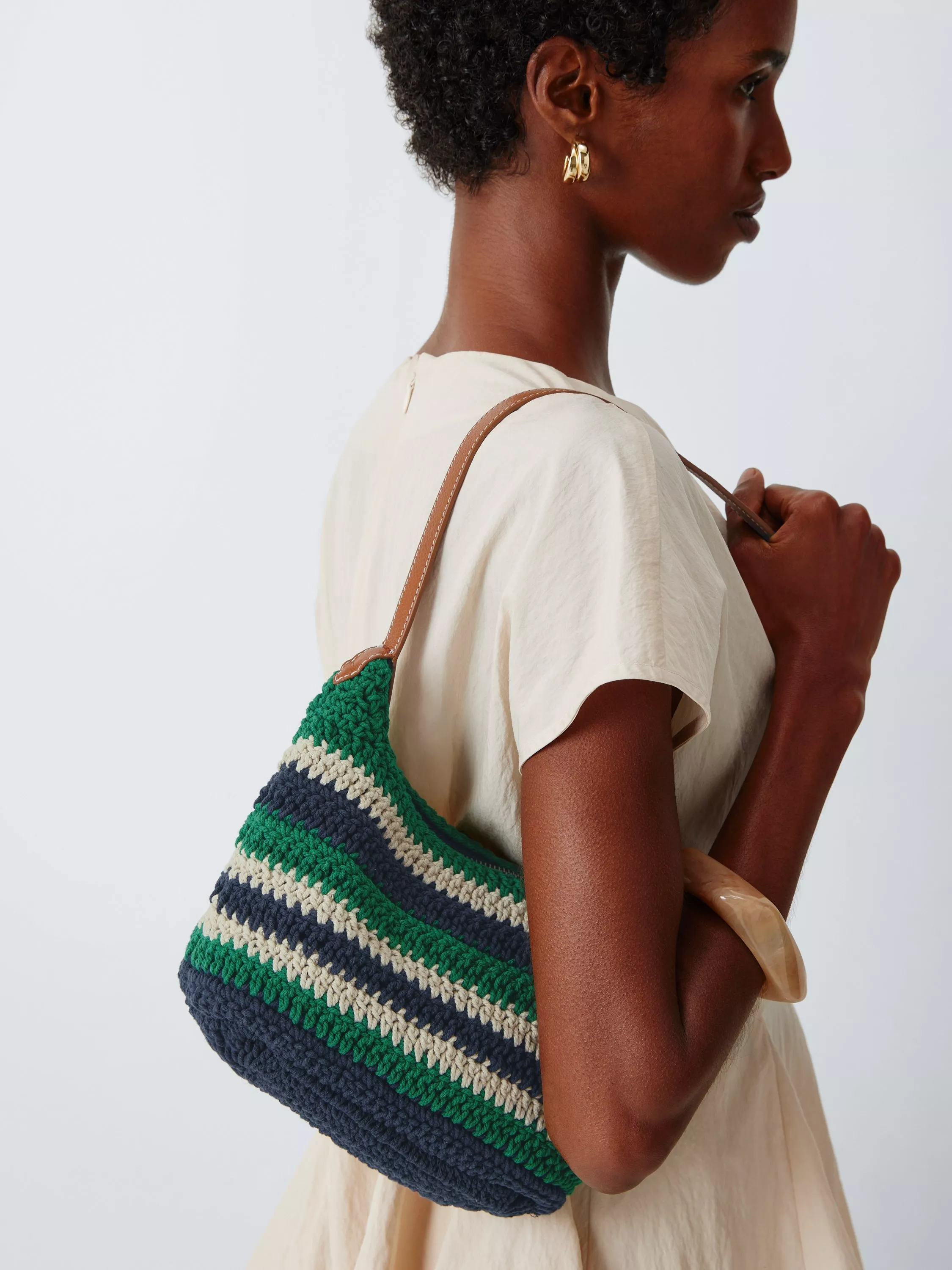 Side profile of woman wearing gold hoops and carrying a striped crochet shoulder bag with leather strap on white dress