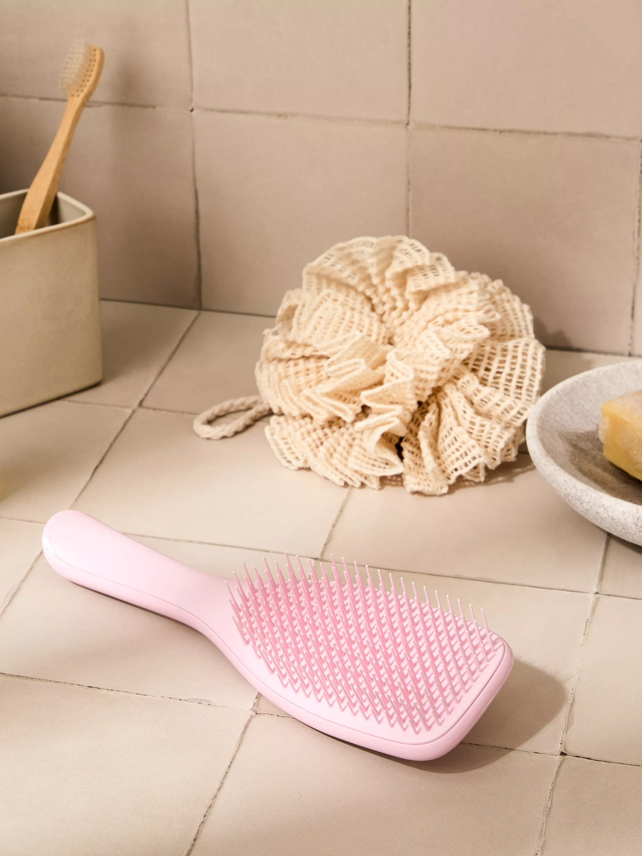 Pink detangling hairbrush on bathroom tiles with bamboo toothbrush, loofah and soap dish in background