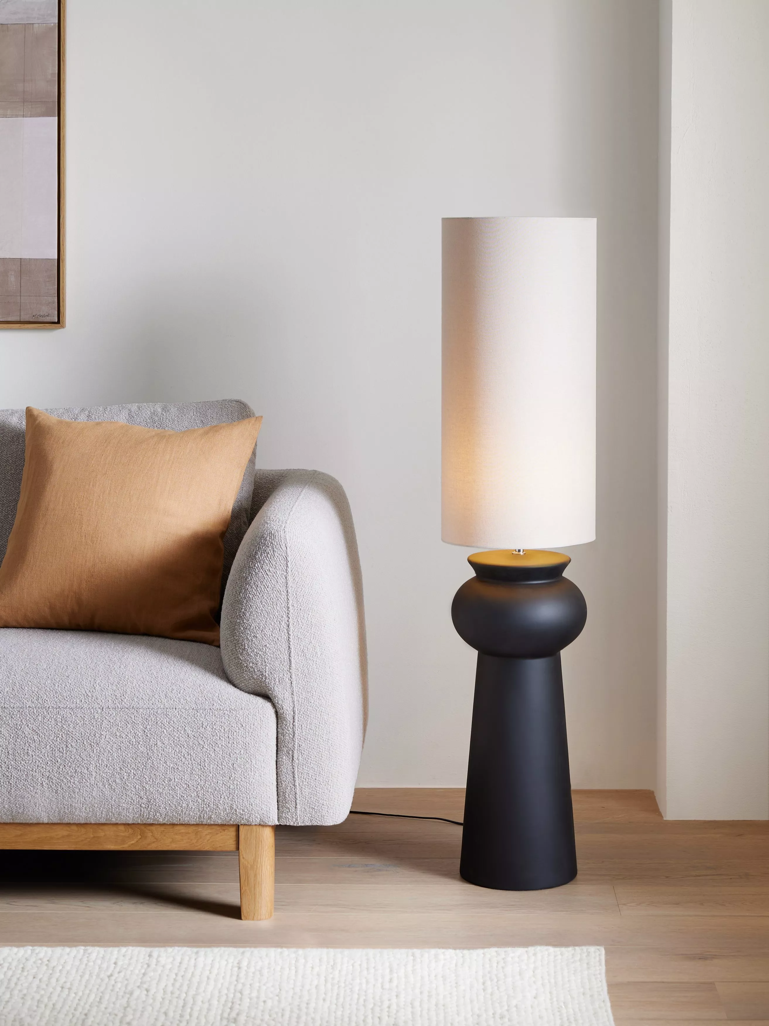 Modern living room corner with grey sofa and tan cushion beside a black ceramic table lamp with white shade