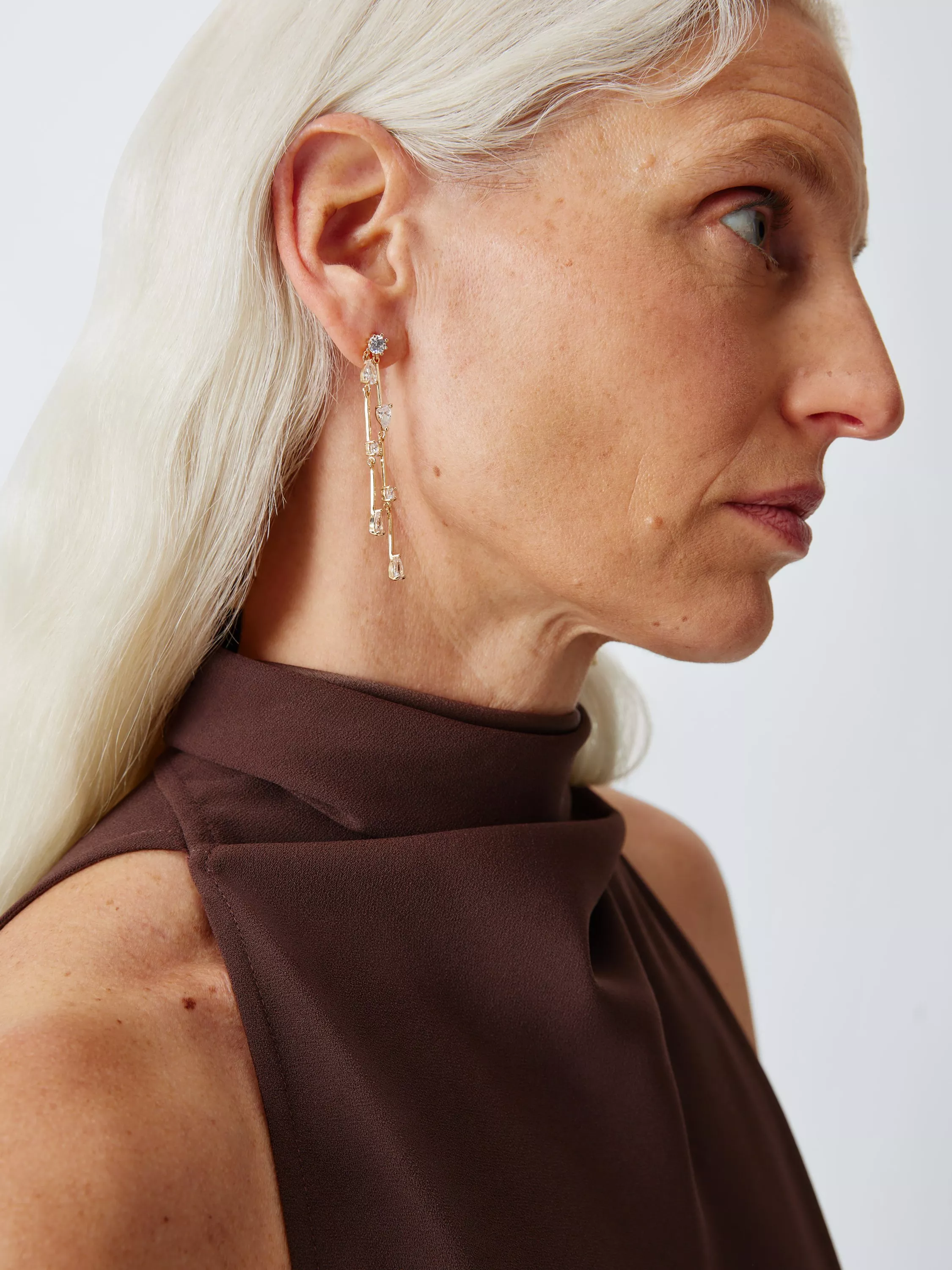 Woman with long white hair wearing a dangling earring and a brown sleeveless top, gazing sideways.