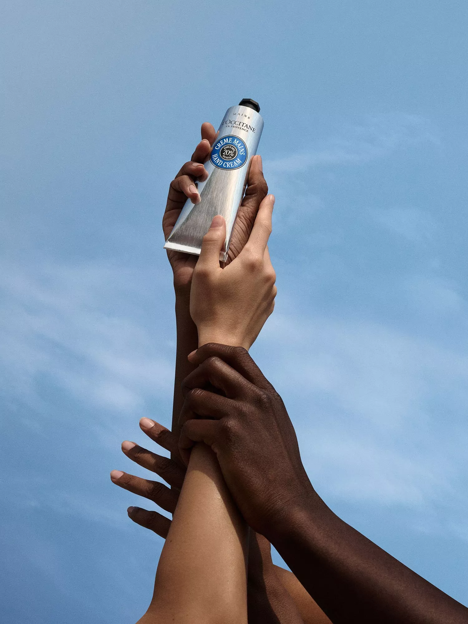 Diverse hands holding L’Occitane 20% shea butter hand cream tube against blue sky, skincare product shot