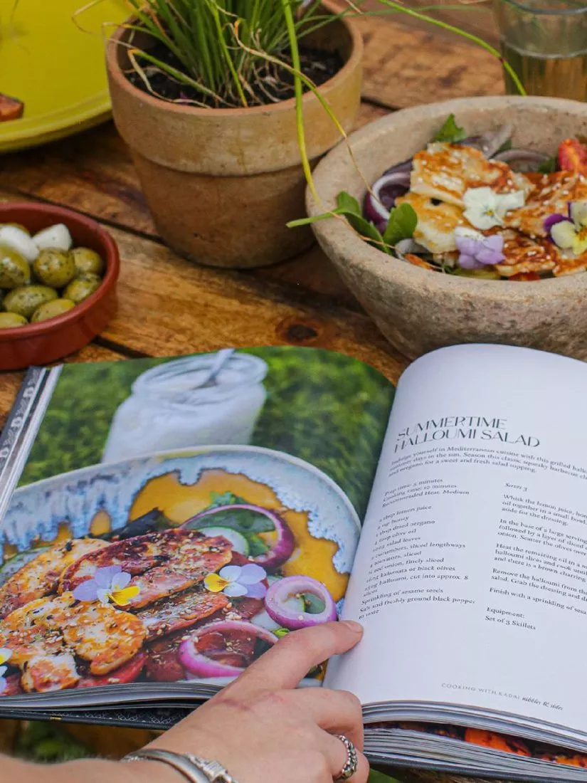 Open cookbook showing summertime halloumi salad recipe on rustic table with olives, potted herbs and finished dish