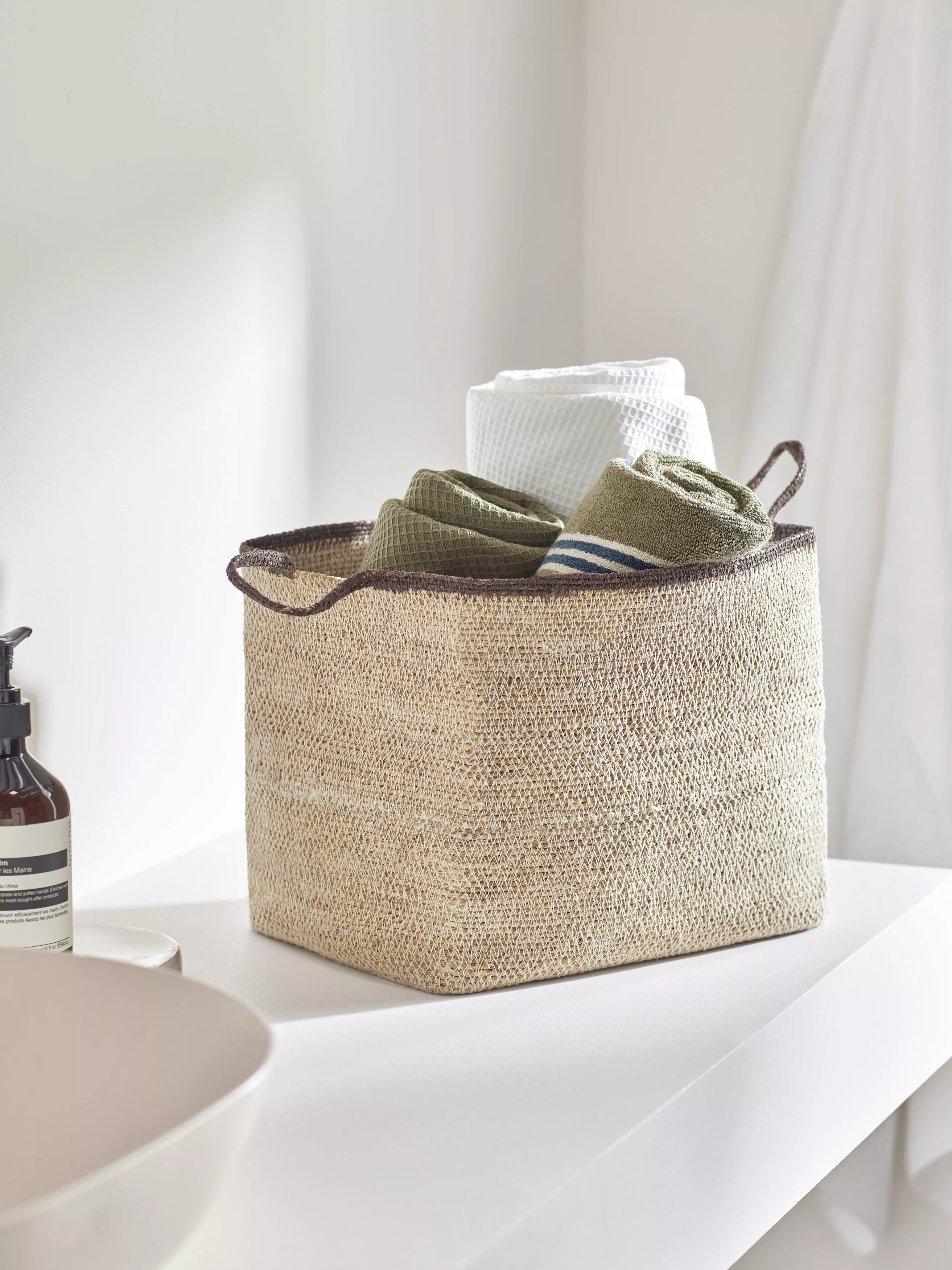 Woven seagrass storage basket with rolled towels on a white bathroom counter, minimalist home organisation decor