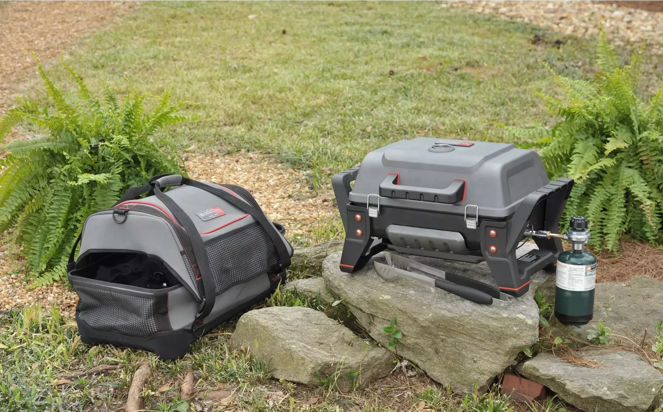 Portable gas barbecue grill with propane canister and carry bag set outdoors on garden stones near ferns