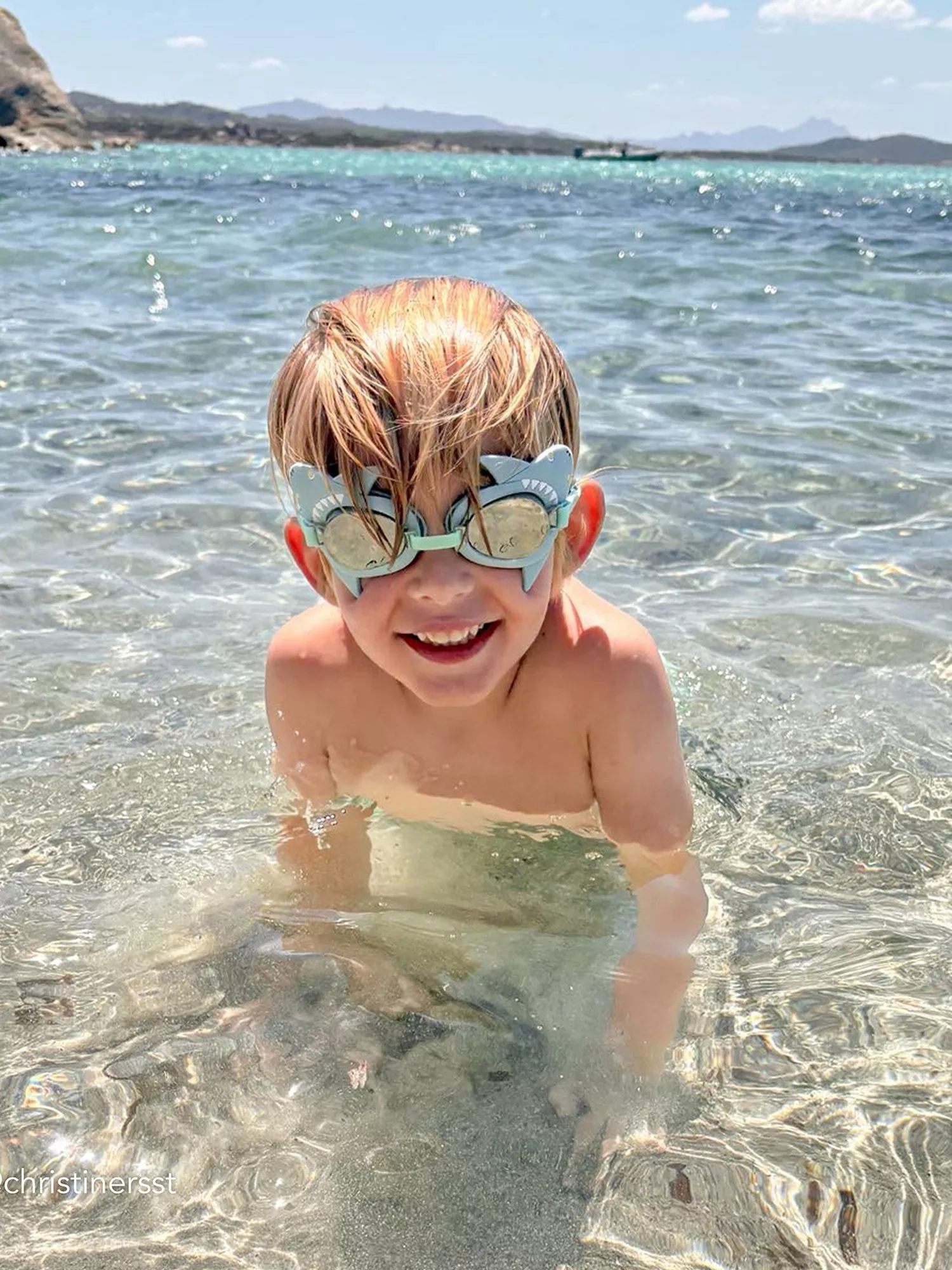 Smiling child wearing shark swim goggles in clear sea water at a sunny beach, with mountains in the distance