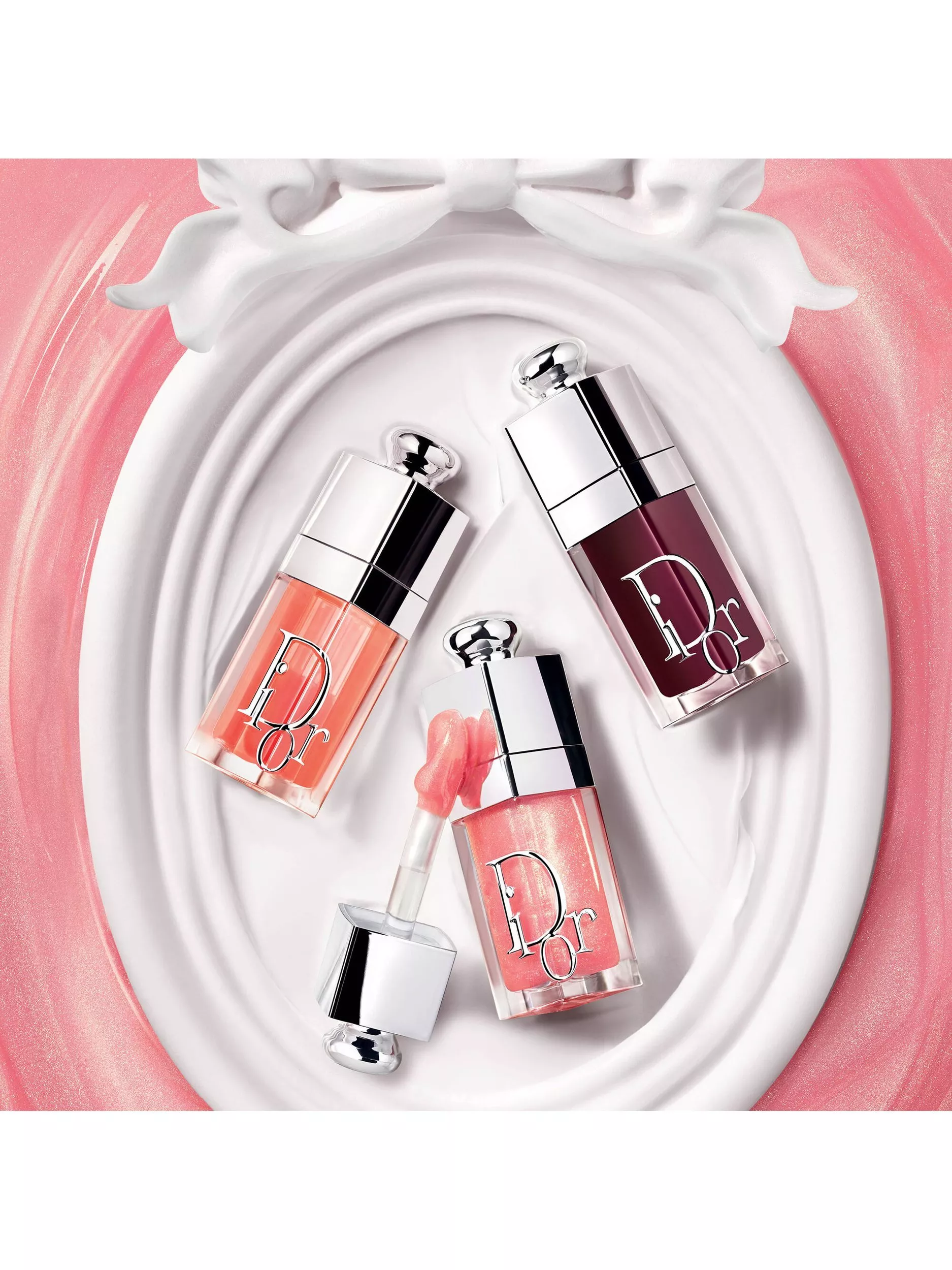 Elegant display of Dior lip glosses in pink and burgundy shades on a decorative white background.