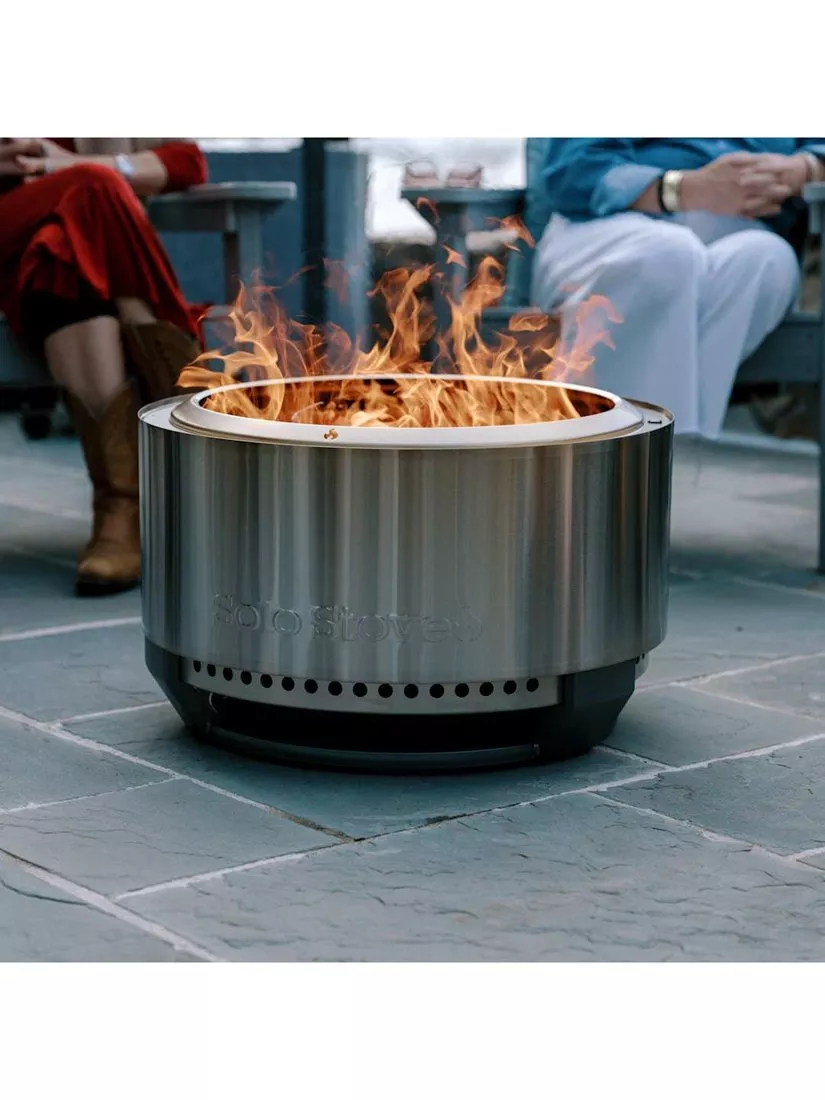 Stainless steel smokeless fire pit burning on patio tiles, with people seated nearby outdoors for warmth