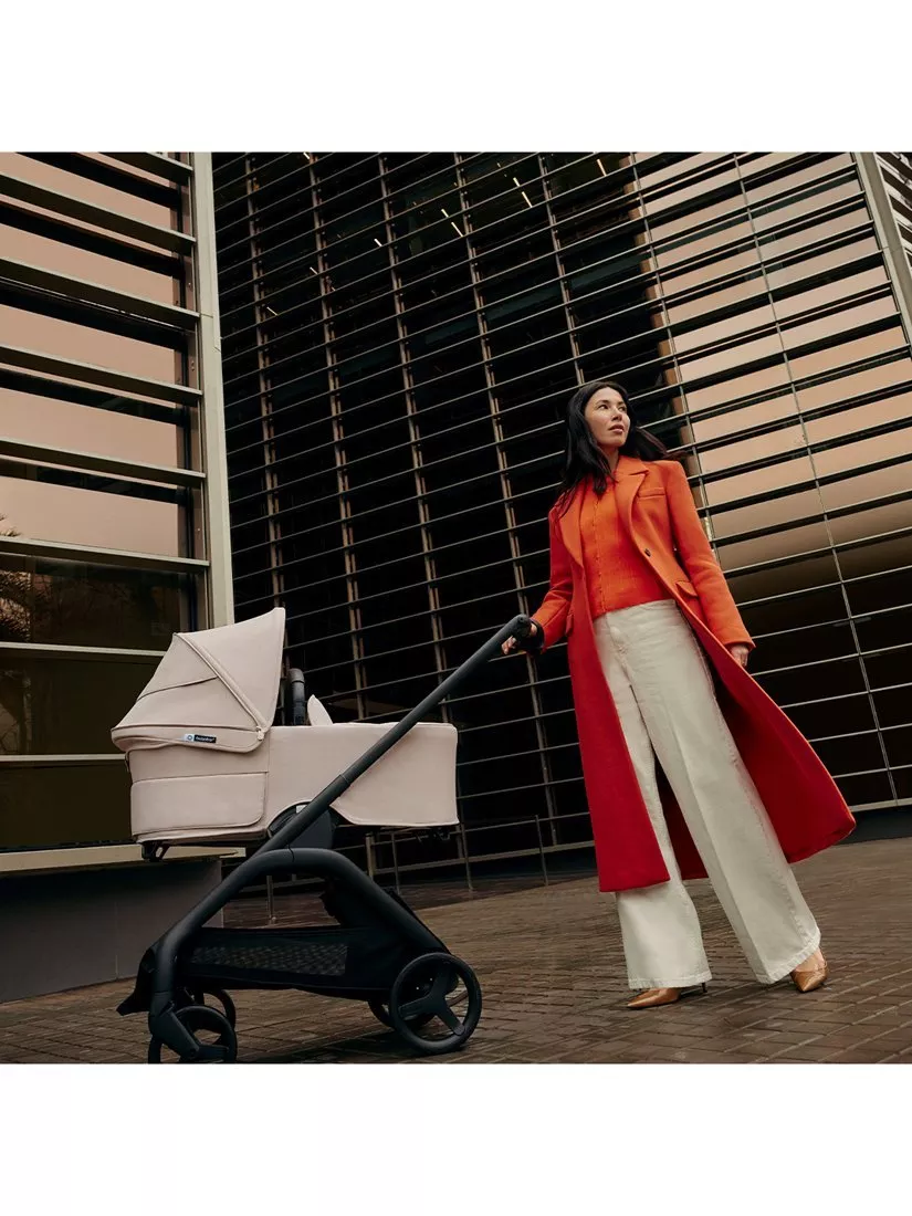 Stylish mother in red coat pushing beige baby pram outside modern building with metal louvres, city street scene