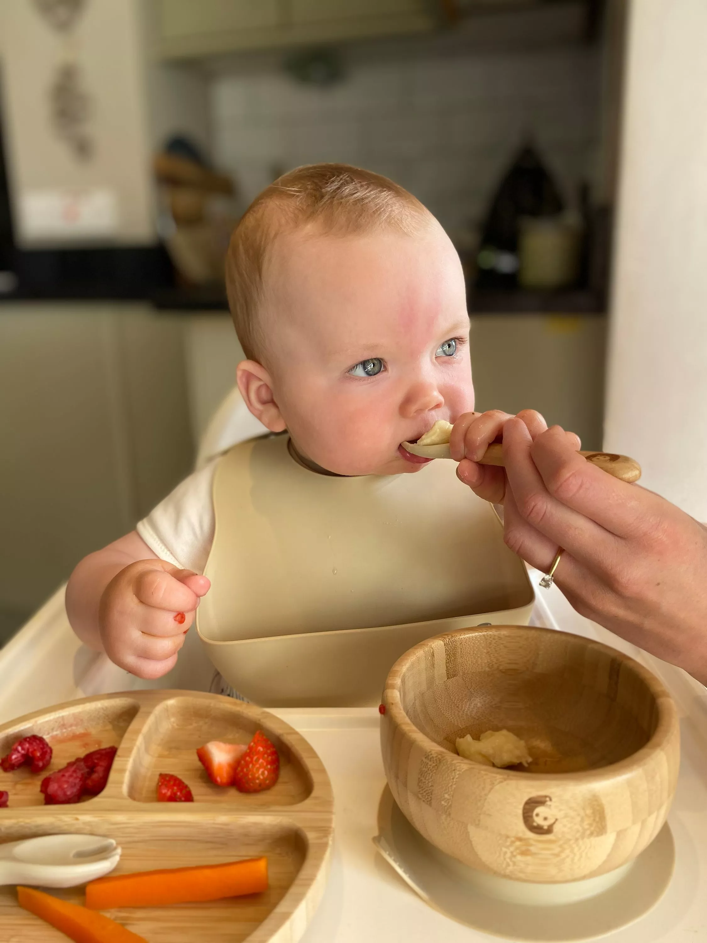 Baby in high chair being spoon-fed puree, with bamboo bowl and fruit on weaning tray in kitchen