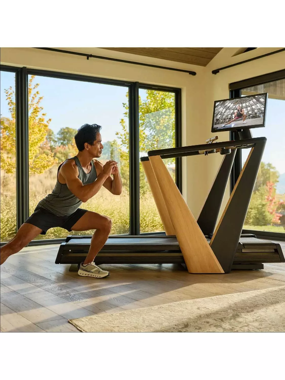 Man doing a lunge workout beside a smart treadmill with screen in a modern home gym with large windows