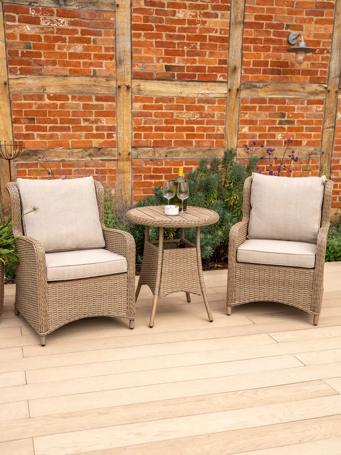 Rattan garden bistro set with two cushioned armchairs and round table on patio, wine glasses by brick wall
