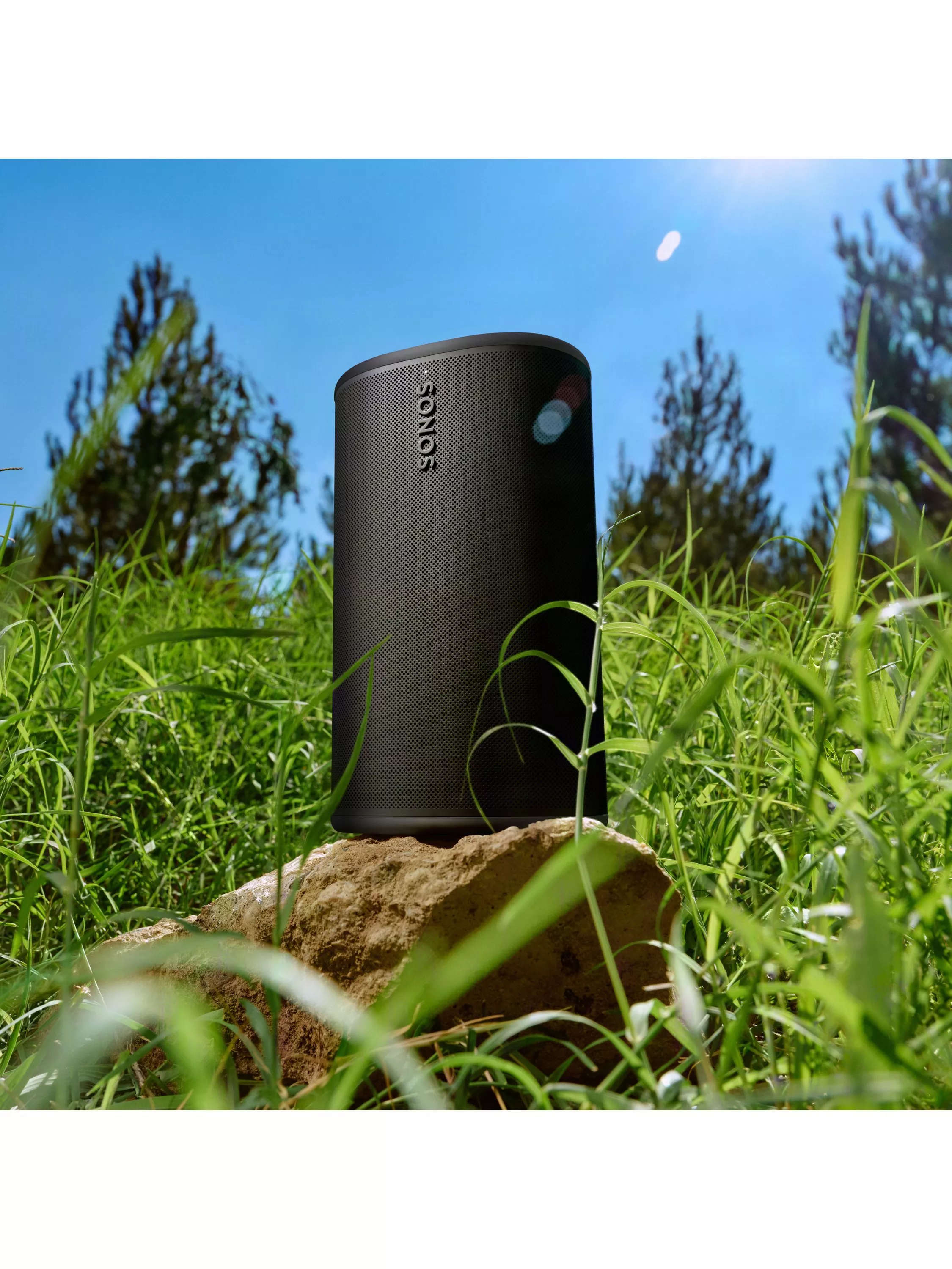 Black Sonos portable Bluetooth speaker on a rock in grassy field under blue sky, outdoor audio lifestyle shot
