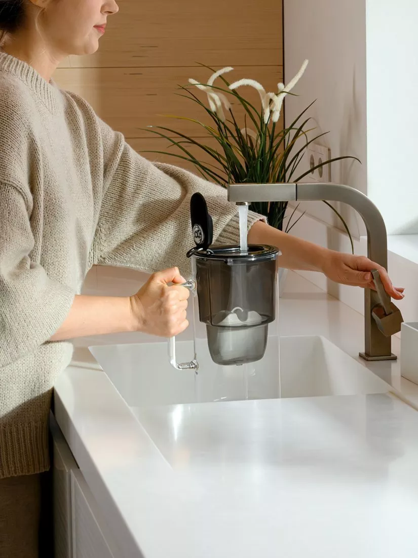 Person filling a water filter jug under a modern kitchen tap at a white sink, home drinking water filtration