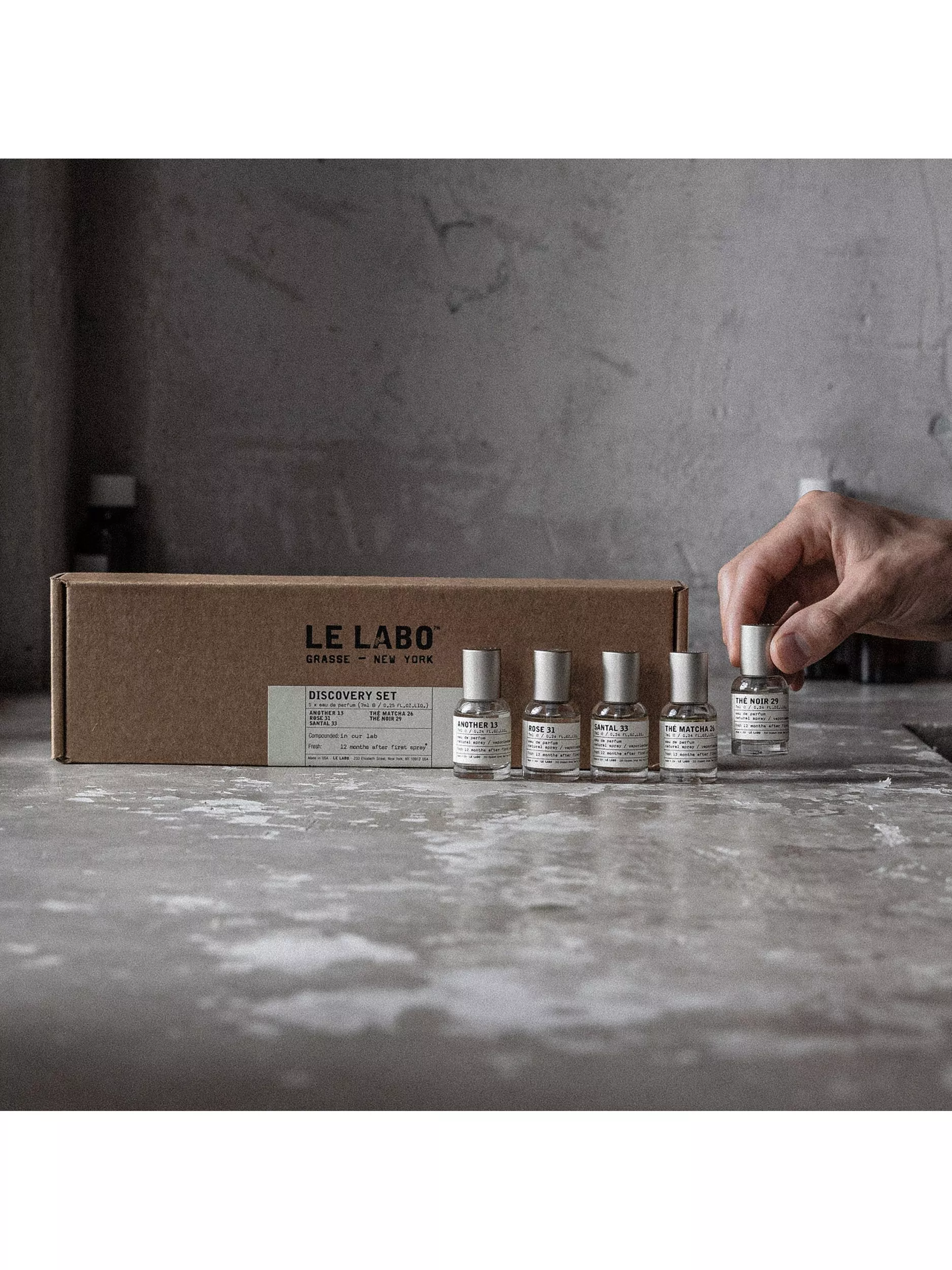 Le Labo Discovery Set perfume samples in front of box, hand holding Thé Noir 29 bottle on concrete surface