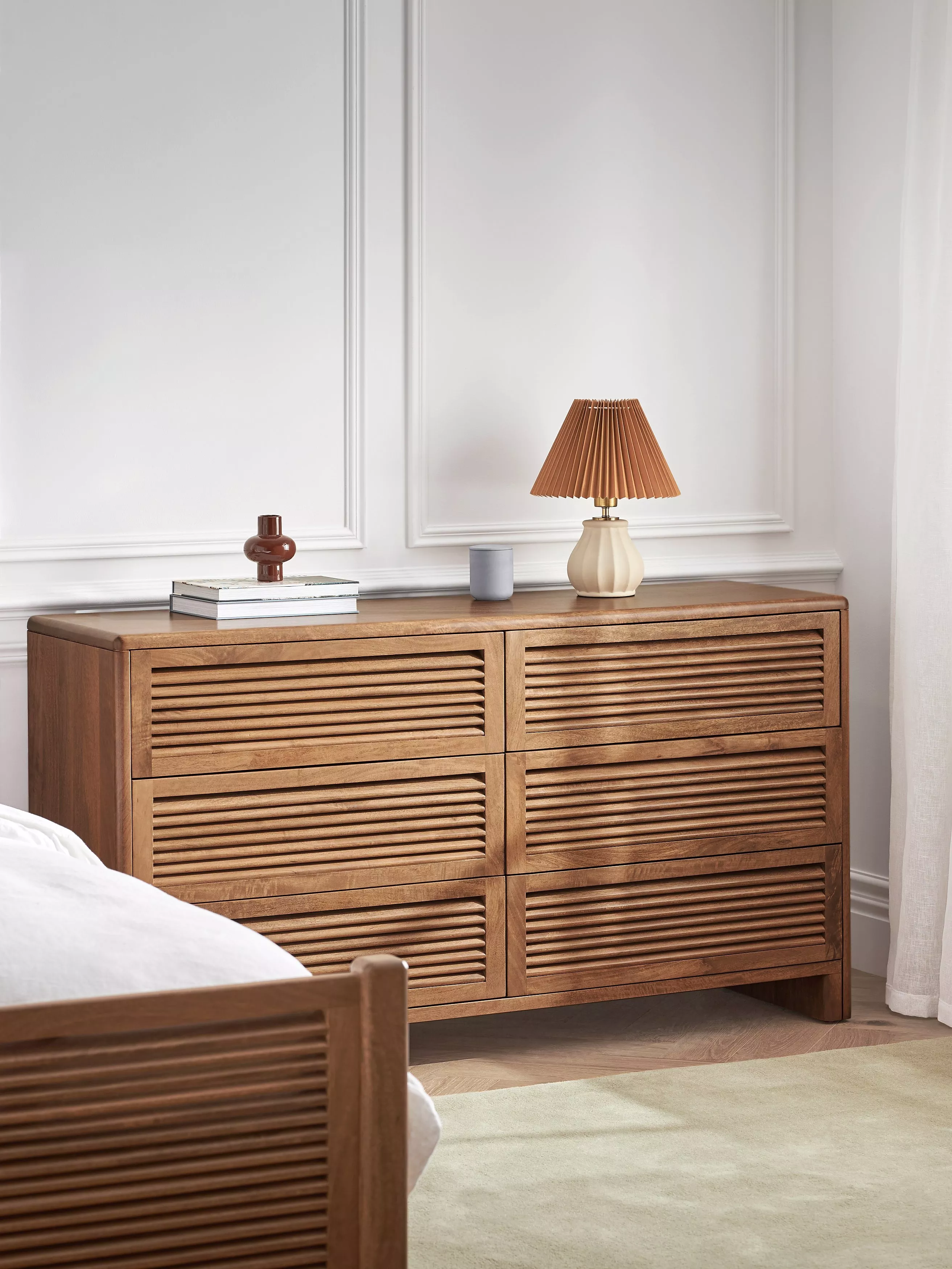 Wooden slatted dresser with six drawers, styled with lamp and books in a modern bedroom interior.