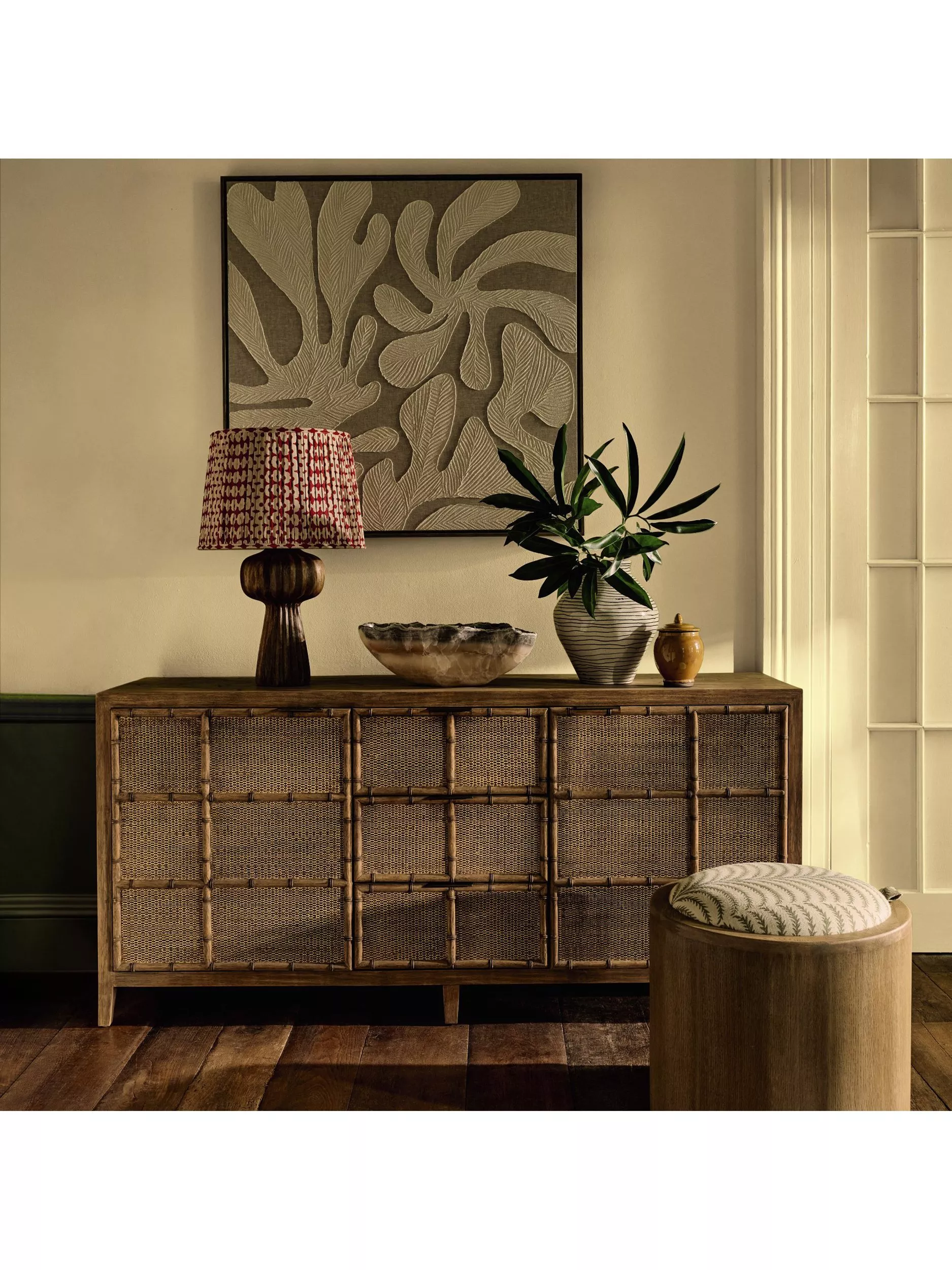 Coastal hallway with rattan sideboard, patterned lamp, leafy plant vase, framed wall art and upholstered stool