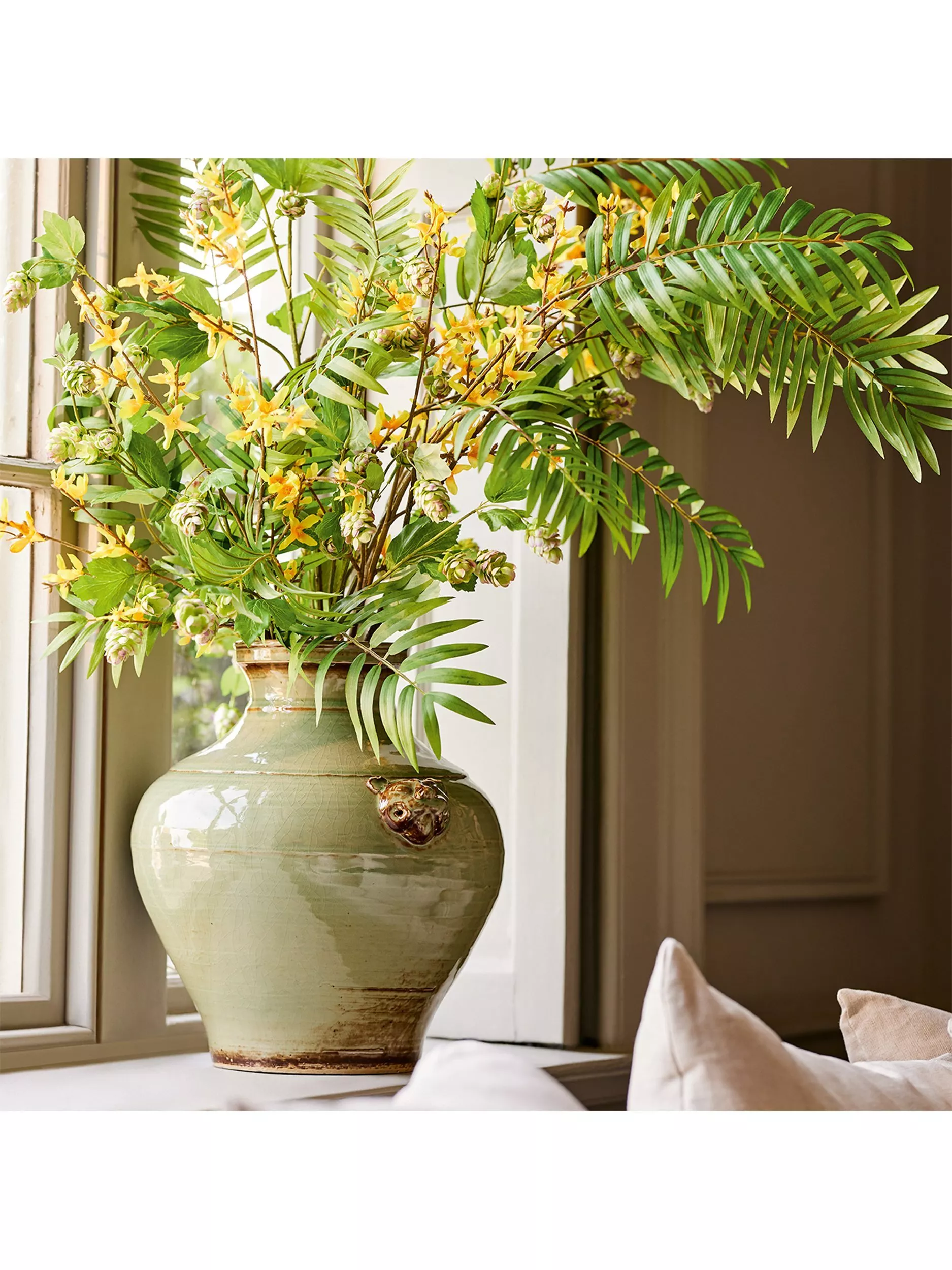 Large ceramic vase on windowsill with lush green foliage and yellow flowers, styled as elegant home décor arrangement